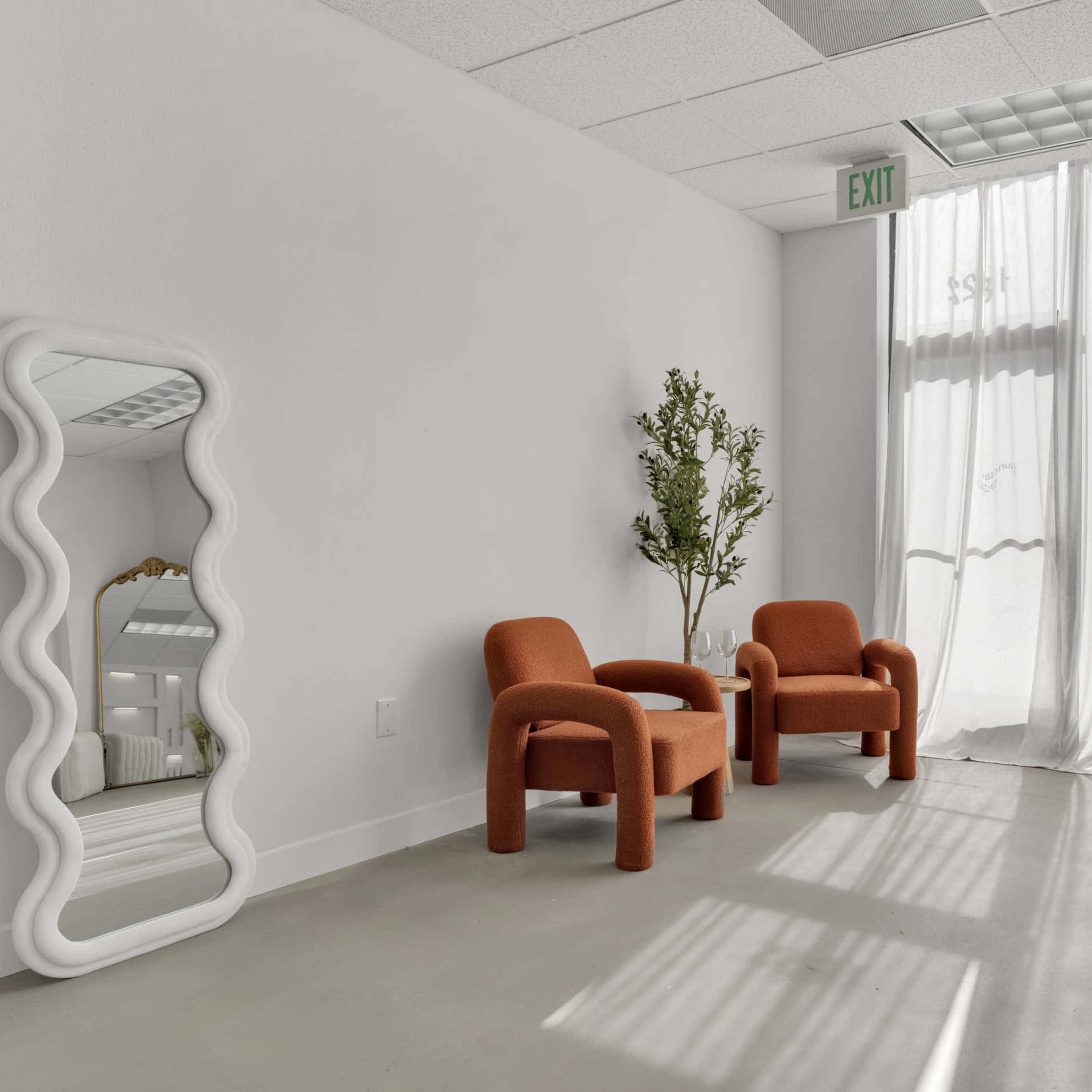 A bright room with two orange chairs, a tall plant, a wavy mirror, and sheer curtains letting light in from a window.