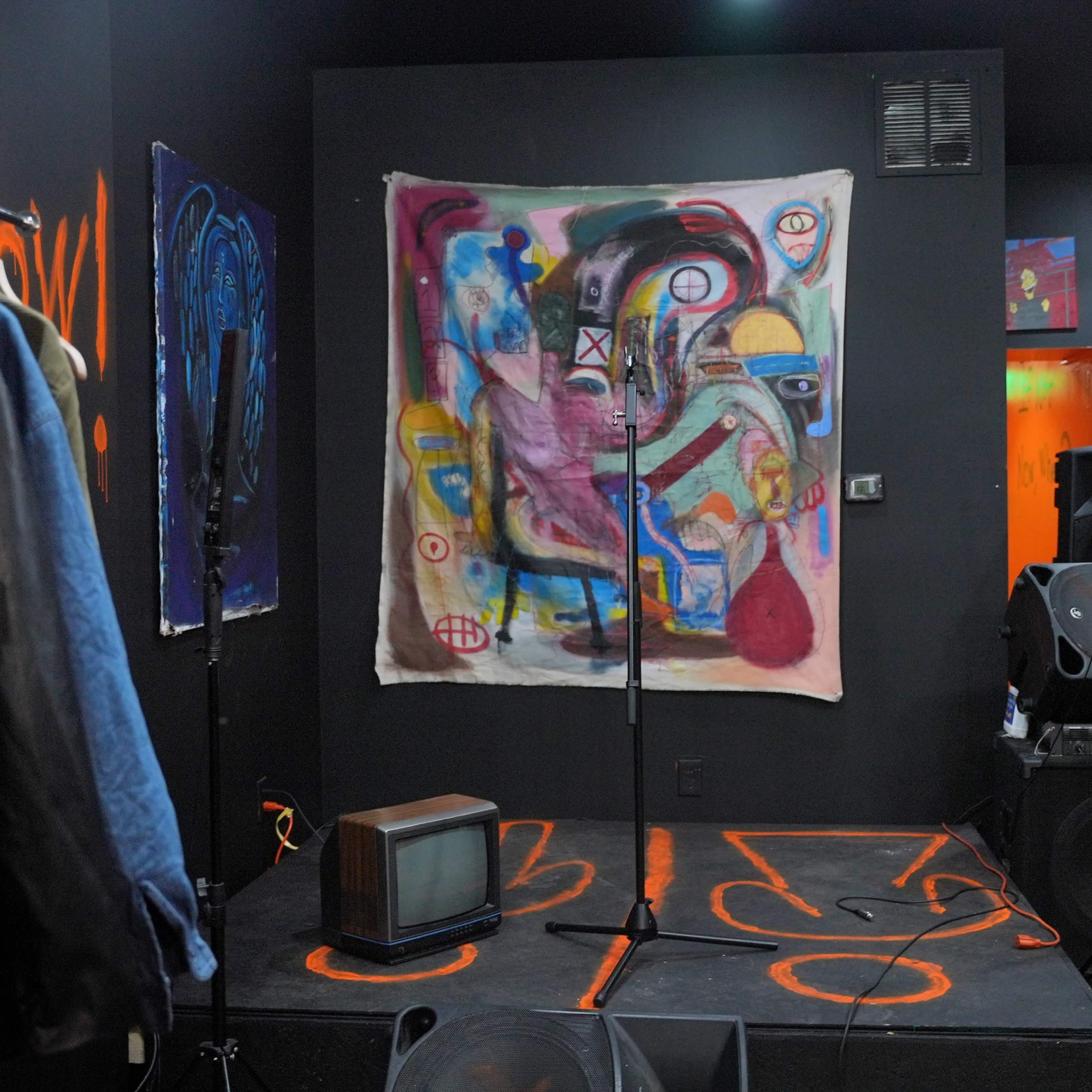 A dark room with a clothing rack, a colorful abstract painting on the wall, and a small stage set up with microphones and an old television.