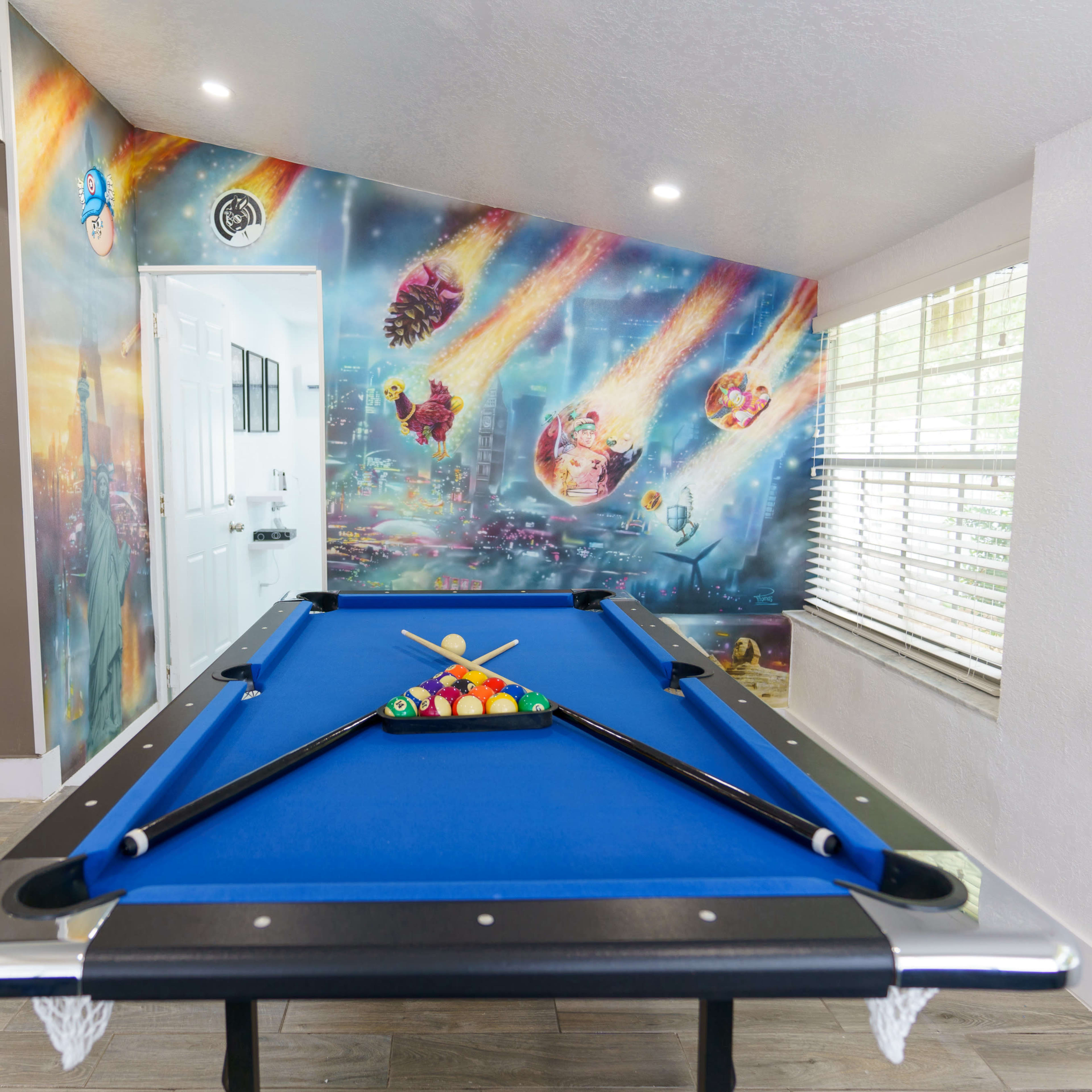 A brightly painted room featuring a blue pool table in the foreground, with a mural depicting colorful space scenes on the wall behind.