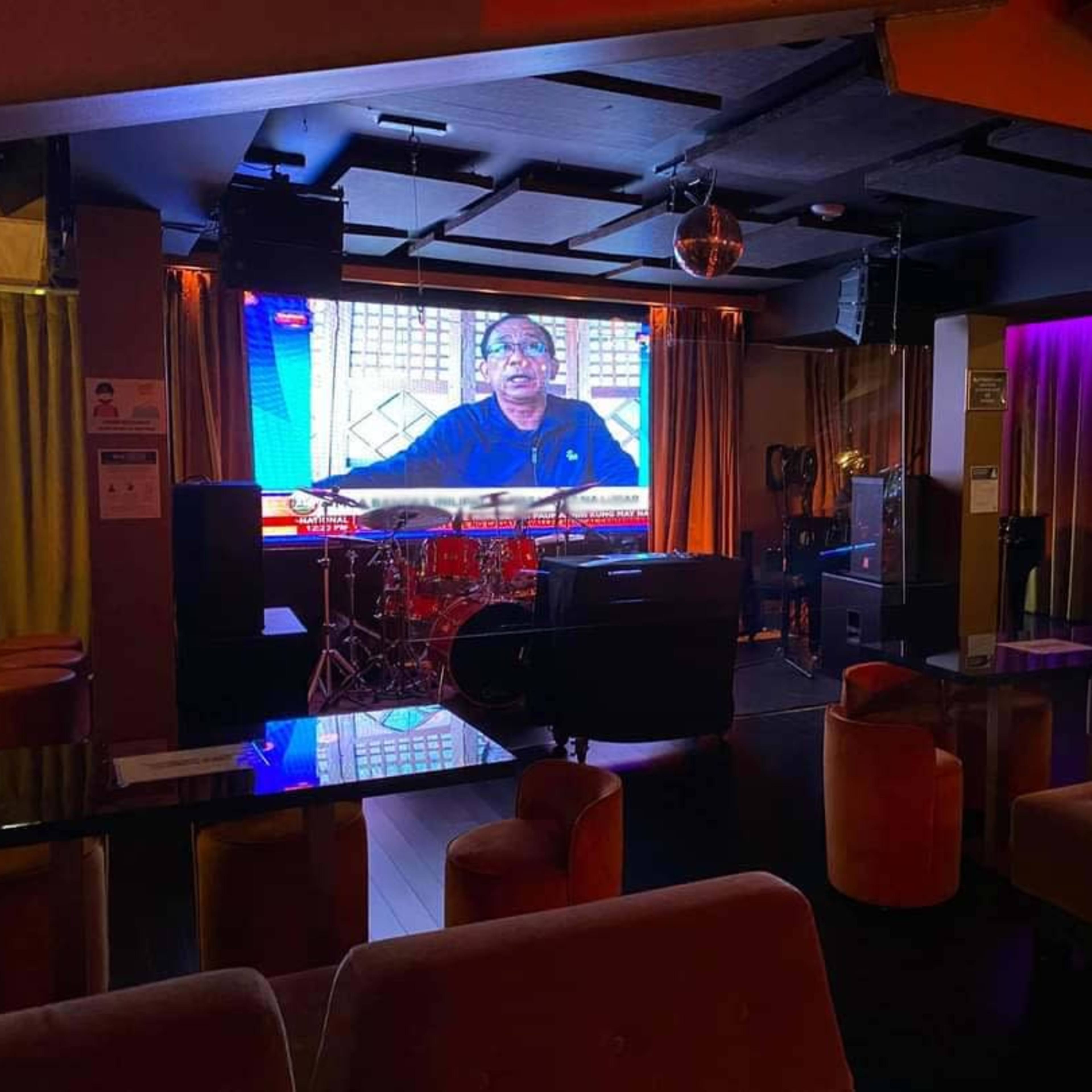 A dimly lit lounge with a large screen displaying a person, surrounded by seating and a small stage with musical instruments.