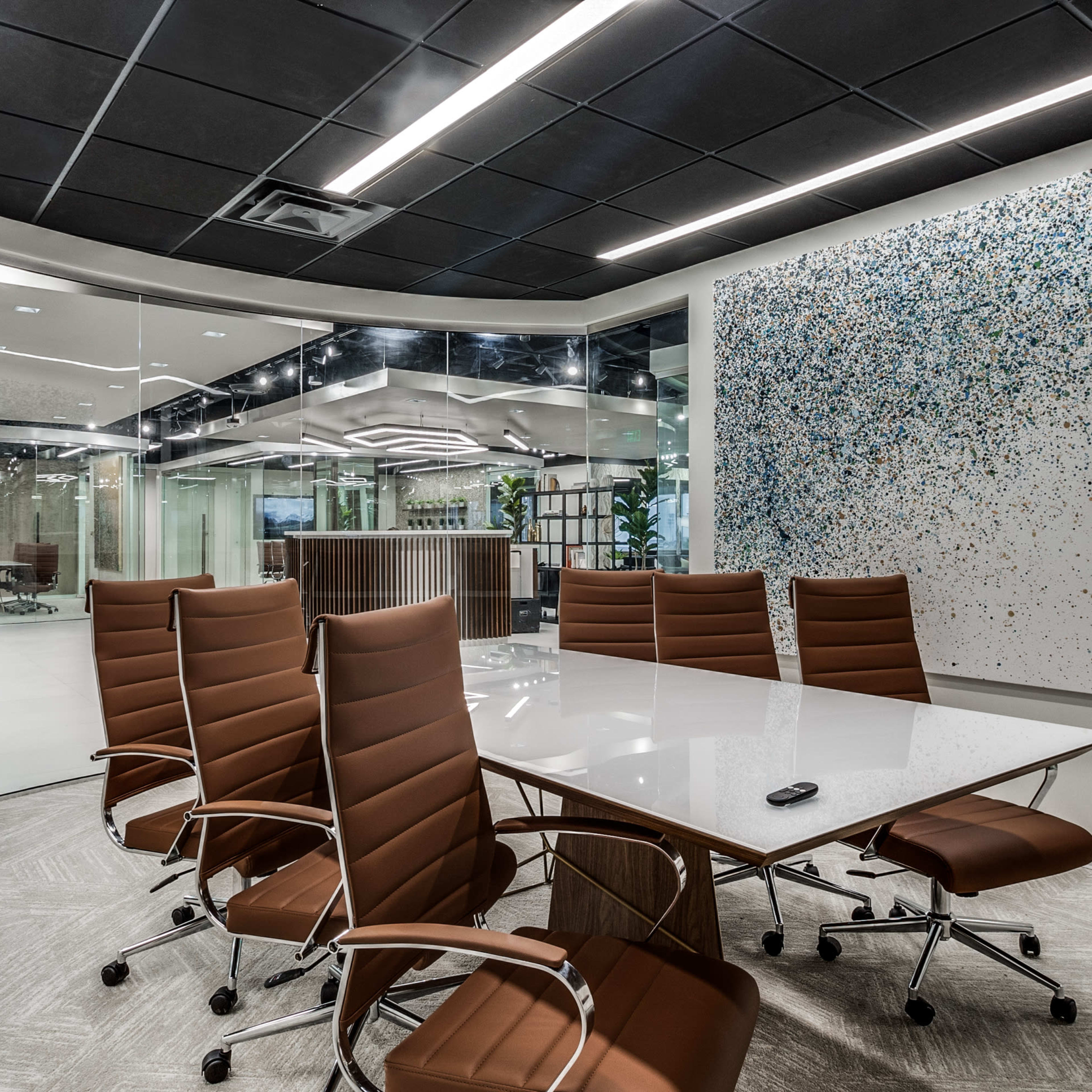 A modern conference room features a large glass table surrounded by six brown chairs, with a decorative wall art piece and glass walls separating the space from adjacent areas.