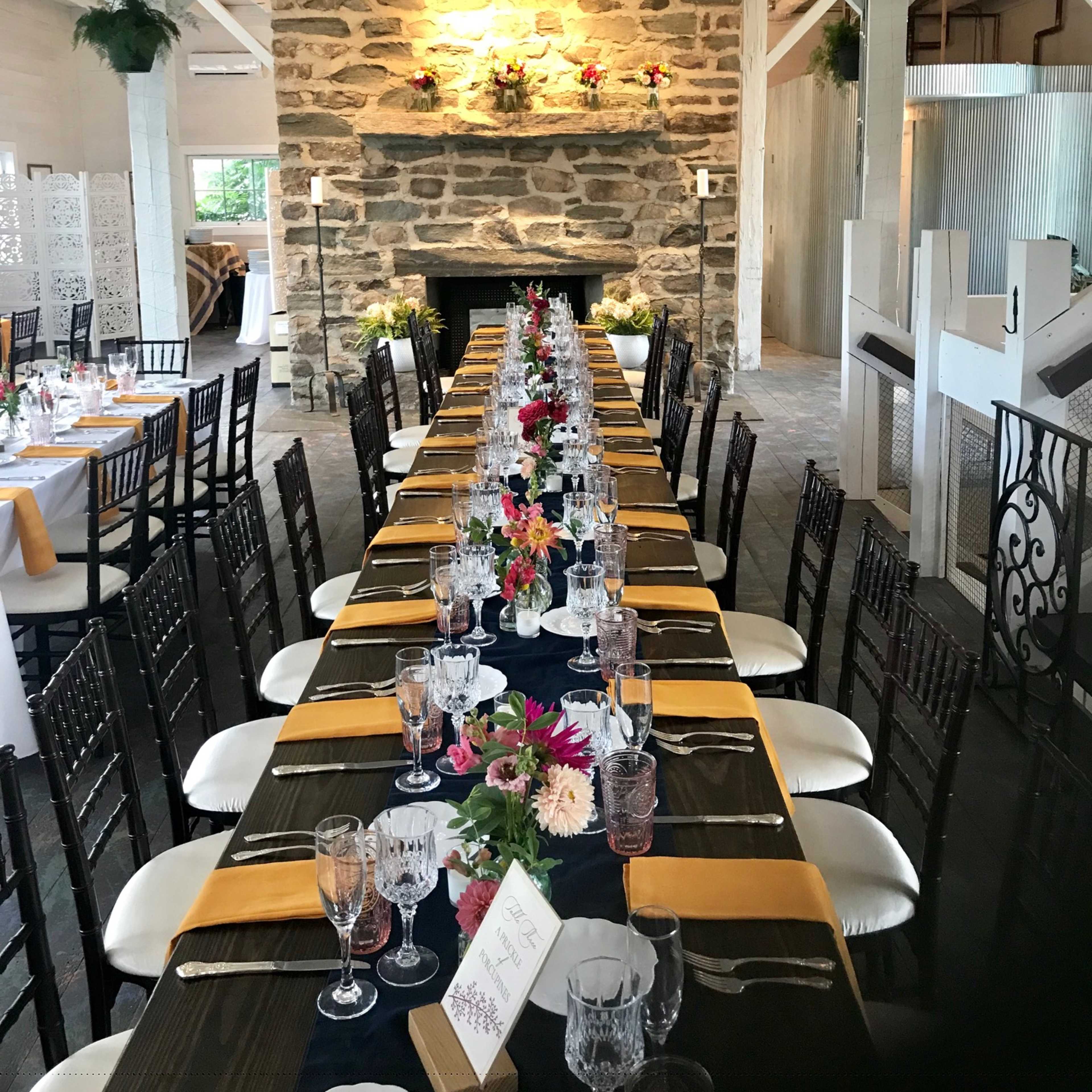 A long banquet table is set with floral centerpieces and dinnerware in a rustic venue featuring a stone fireplace and wooden beams.
