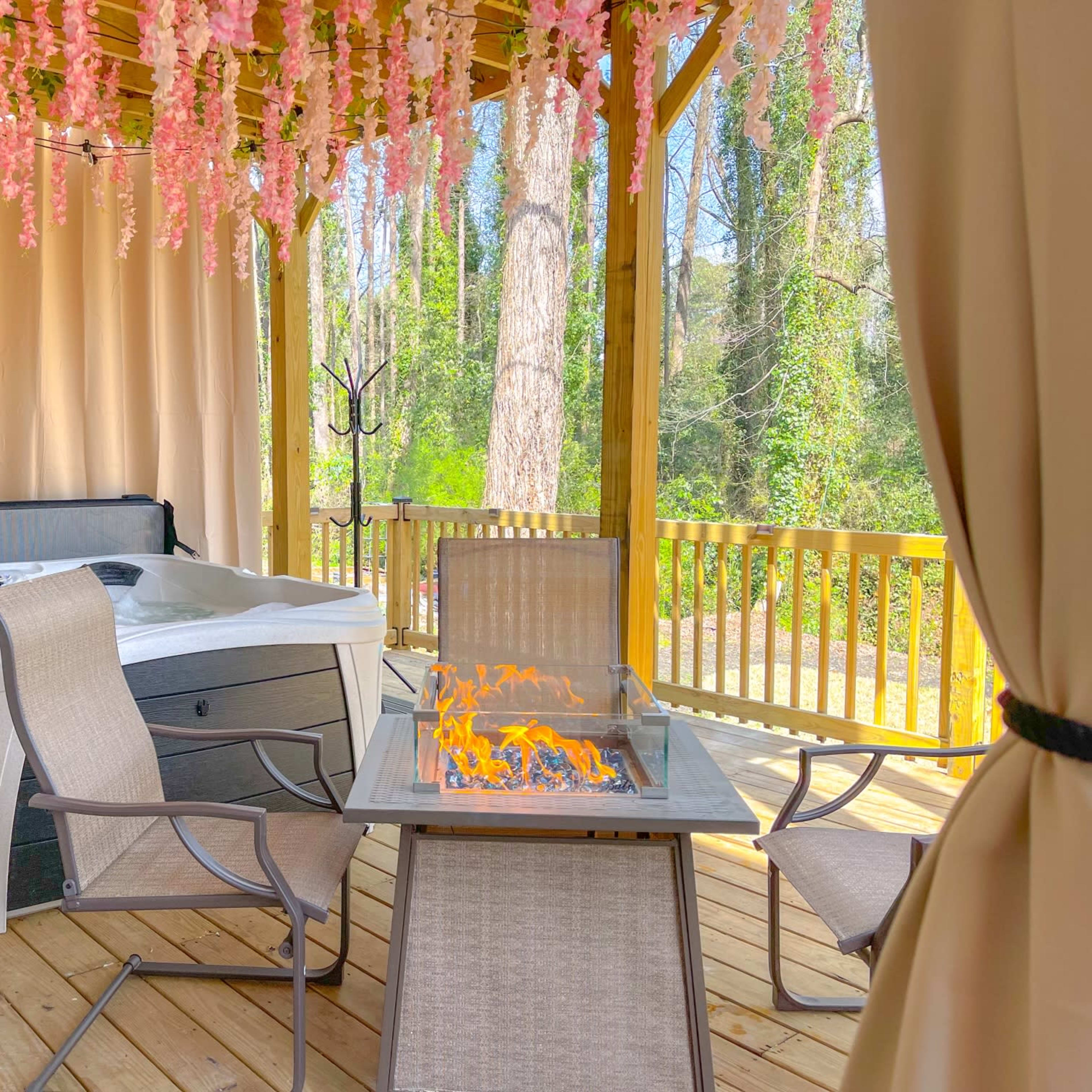 The scene features a hot tub under a canopy decorated with hanging flowers, accompanied by a fire table and several chairs on a wooden deck.