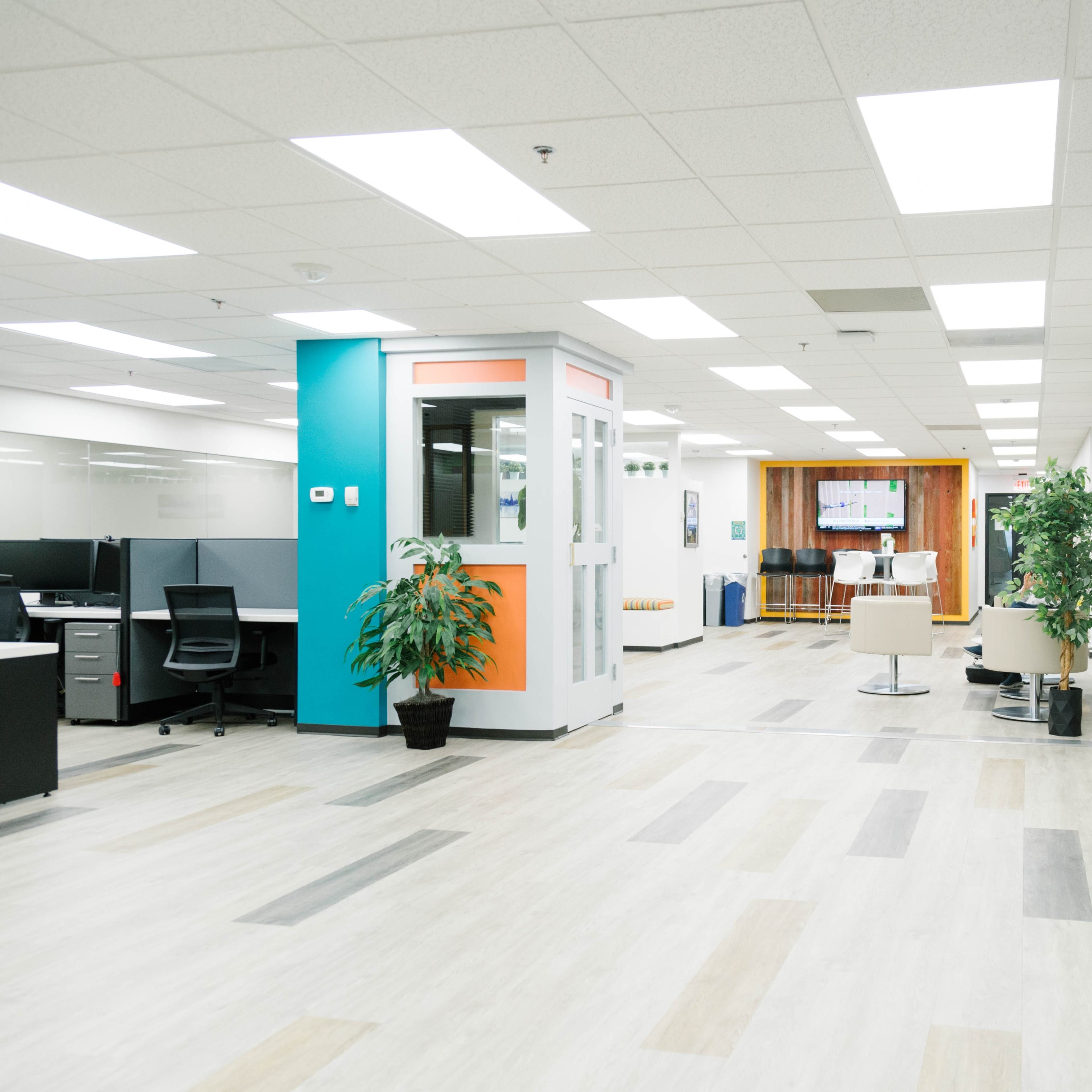 A modern office corridor featuring shared workspaces, a glass-walled meeting room, and plants arranged throughout.