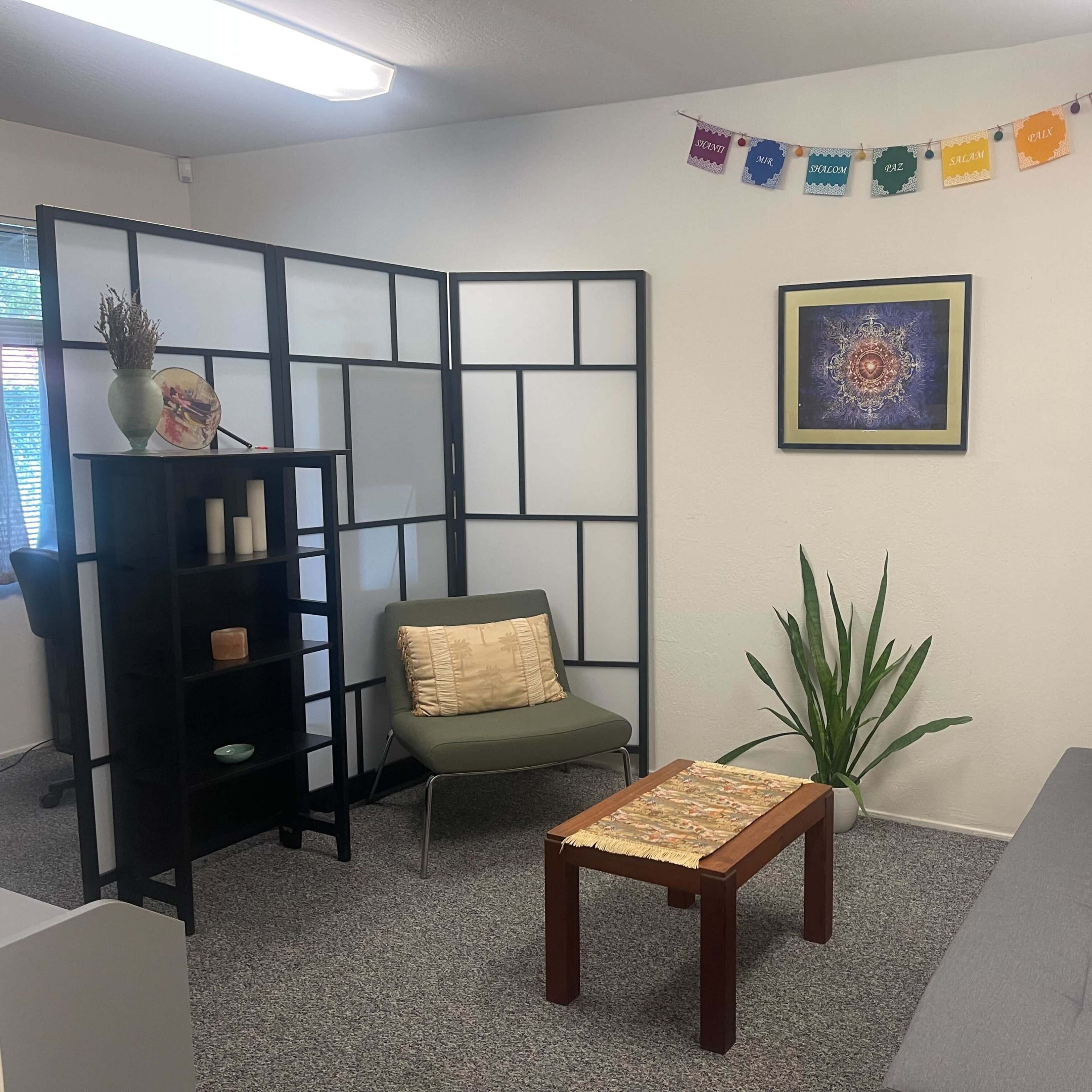 A cozy room with a gray sofa, a wooden coffee table, a plant, a shelf with decorative items, and a colorful banner hanging on the wall.