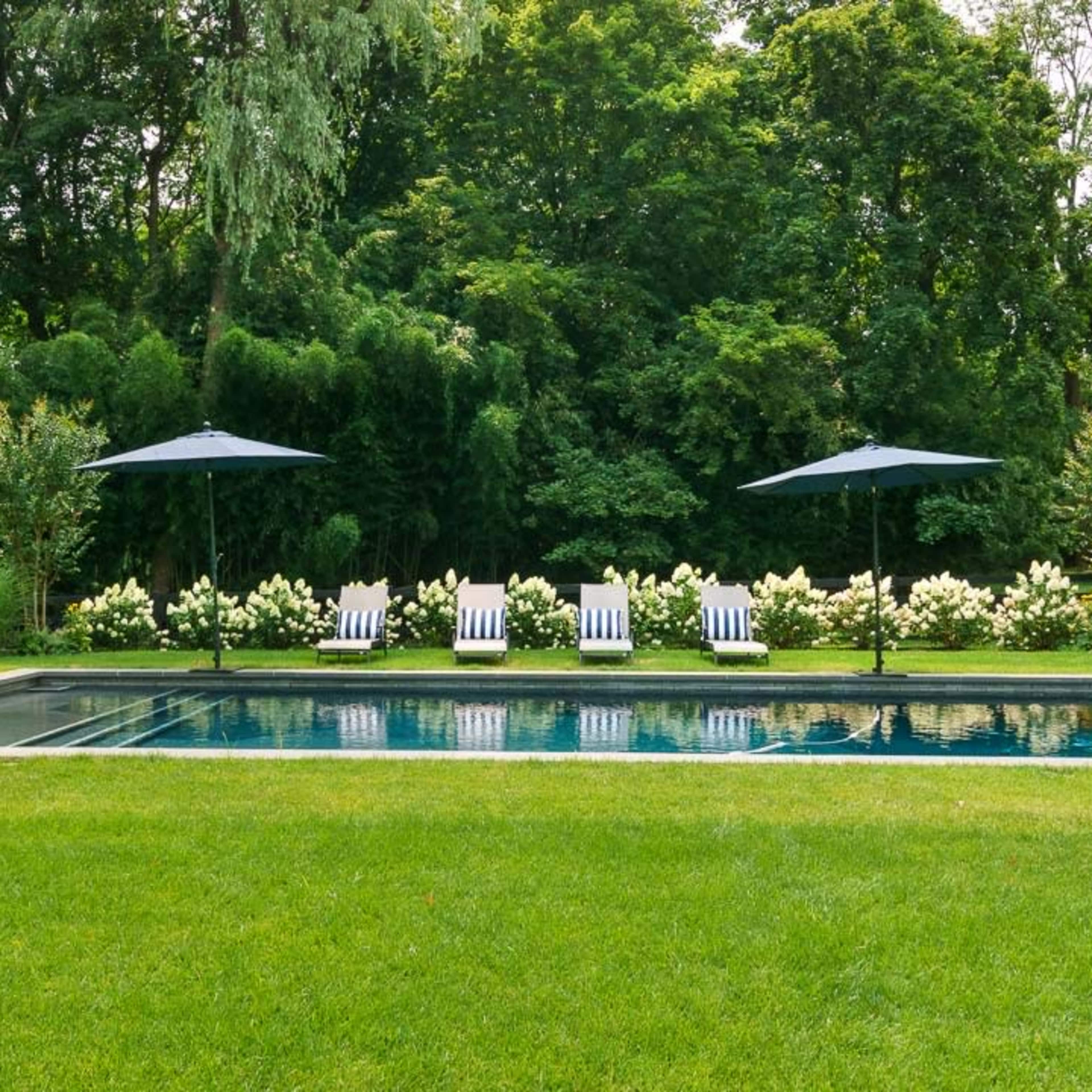 A rectangular swimming pool surrounded by a well-maintained lawn, with white lounge chairs and umbrellas arranged on one side.