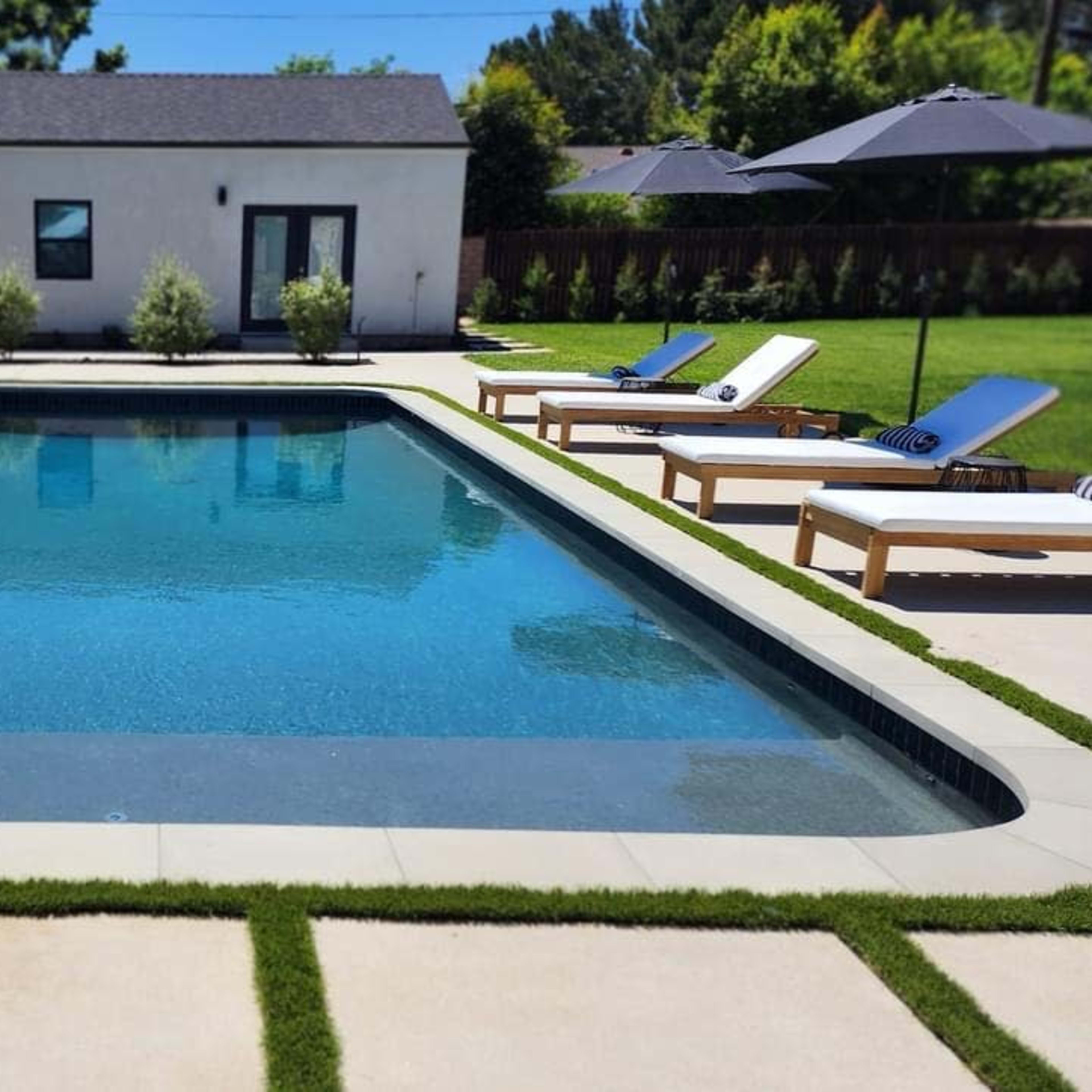 A clear swimming pool is surrounded by lounge chairs on a concrete patio, with a garden area and a small building in the background.