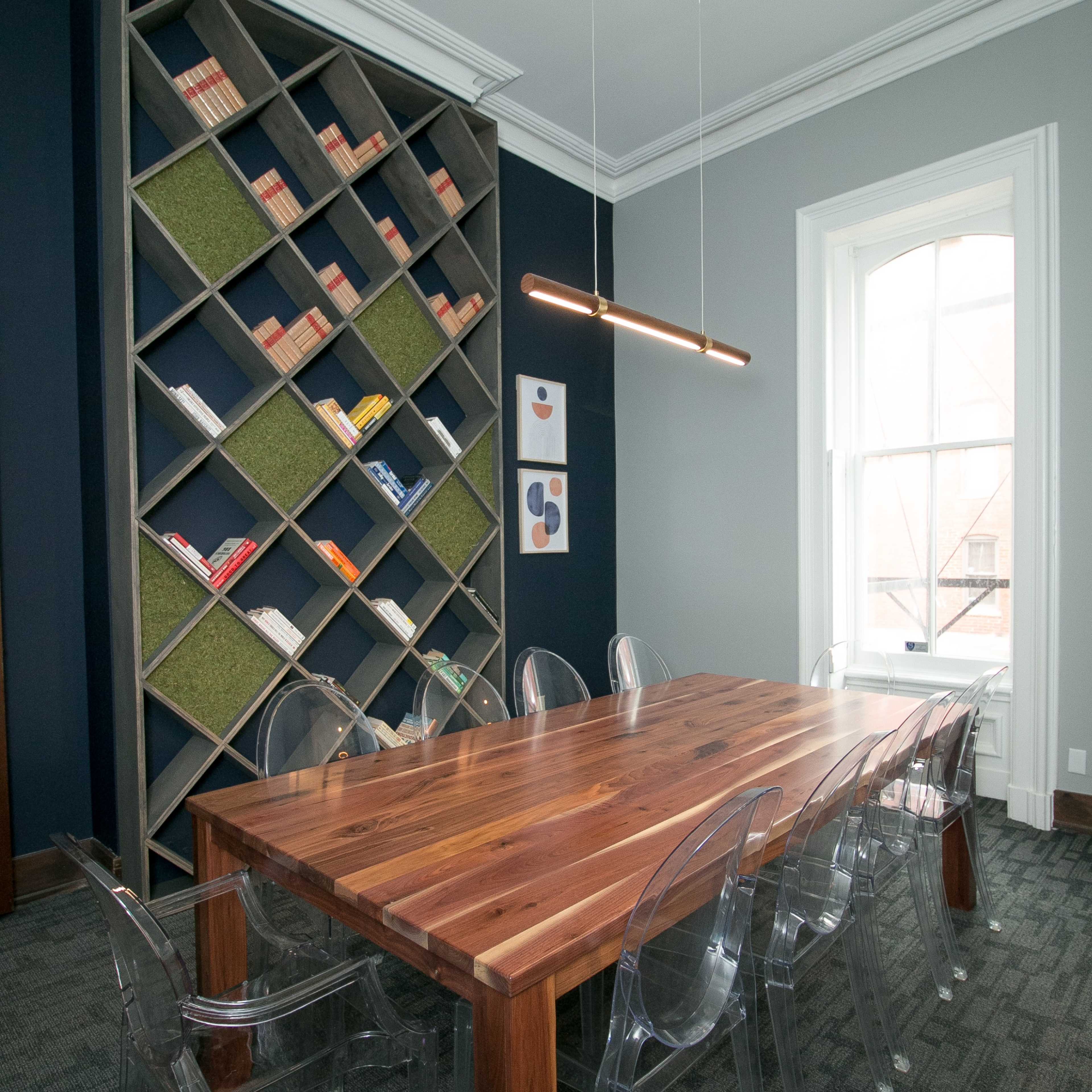 A modern meeting room featuring a large wooden table surrounded by transparent chairs, with a geometric bookshelf filled with books and decorative elements in the background.