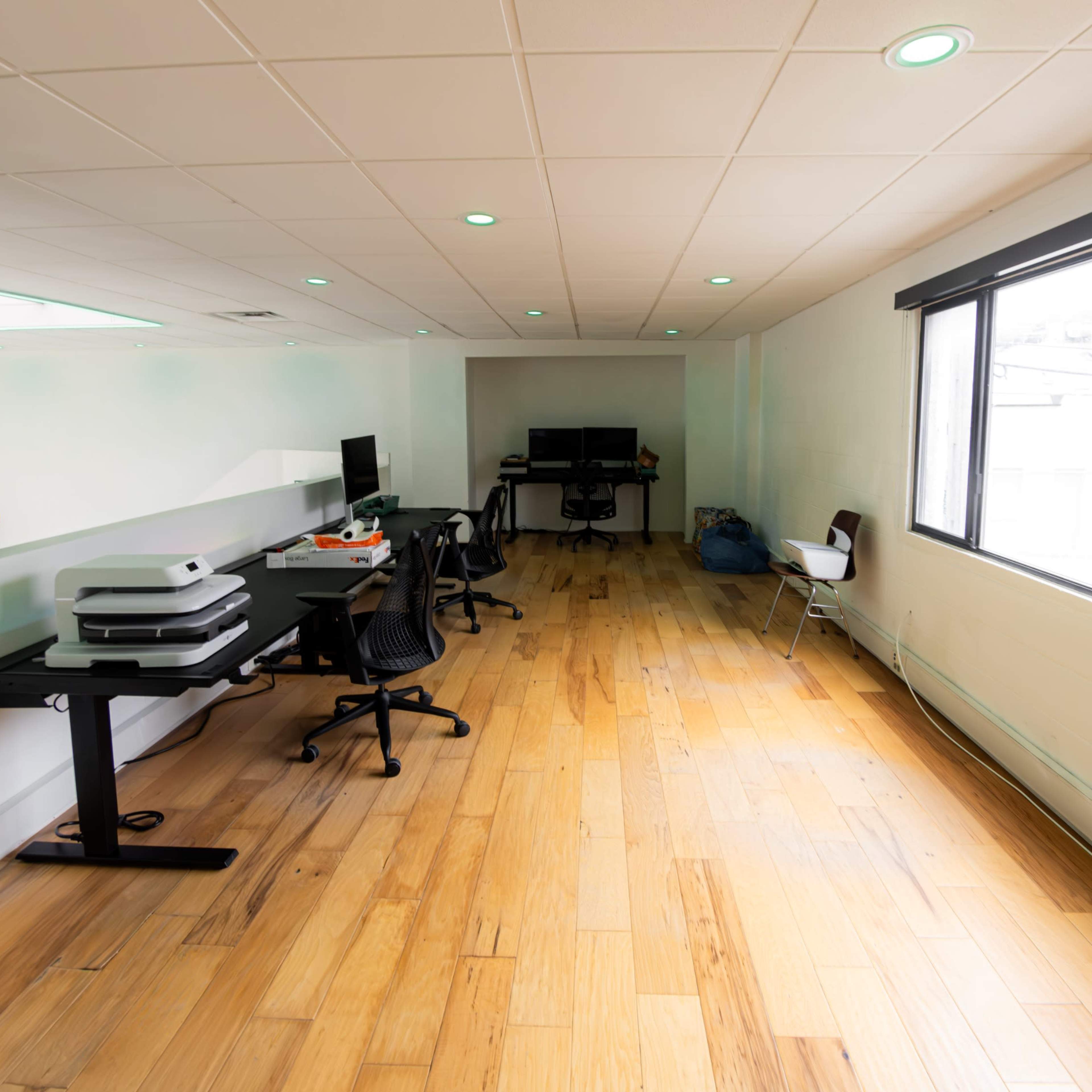 A spacious office area with wooden flooring, equipped with multiple desks, computers, and a large window allowing natural light.
