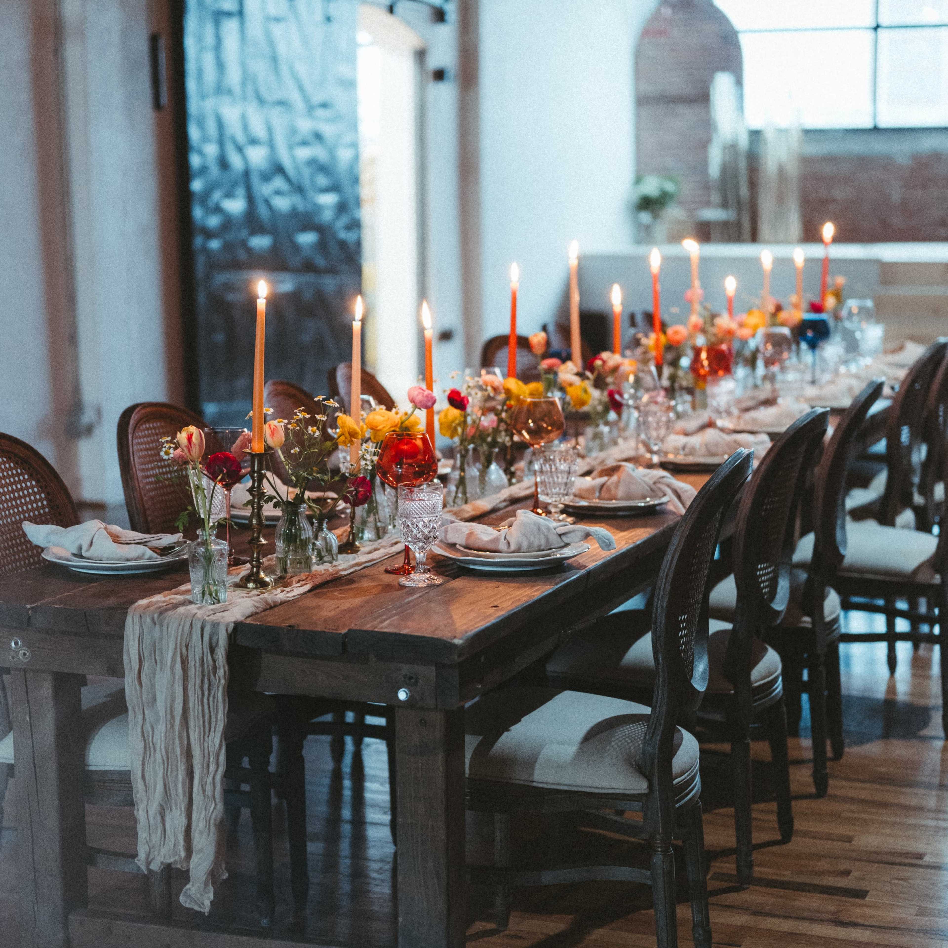 A long wooden table is elegantly set with candles, floral arrangements, and tableware in a well-lit industrial space.