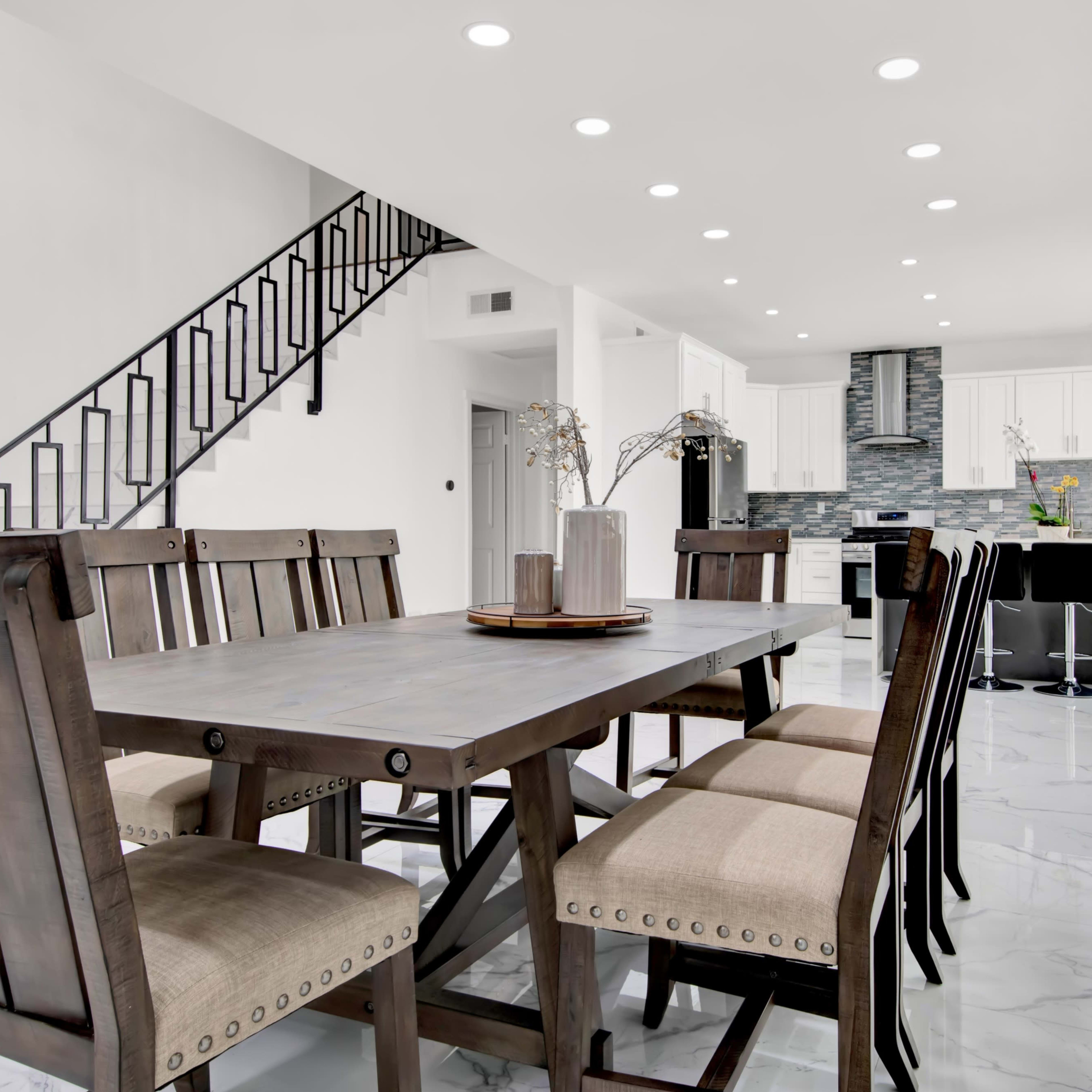 A modern dining area features a wooden table with six chairs and is adjacent to a kitchen with sleek cabinetry and a staircase in the background.
