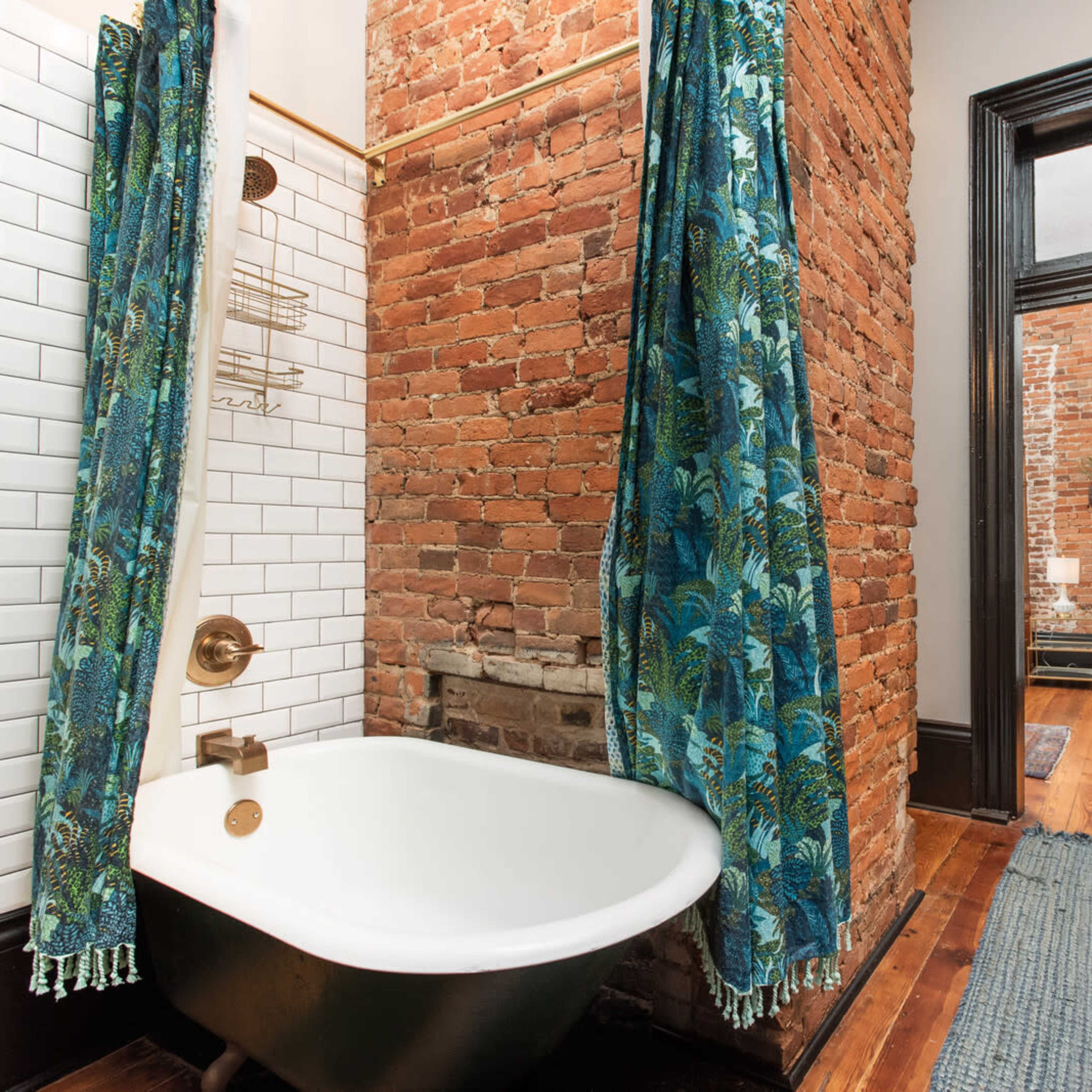 A bathroom with a vintage clawfoot tub, exposed brick walls, and patterned curtains, leading to an adjoining room through a doorway.