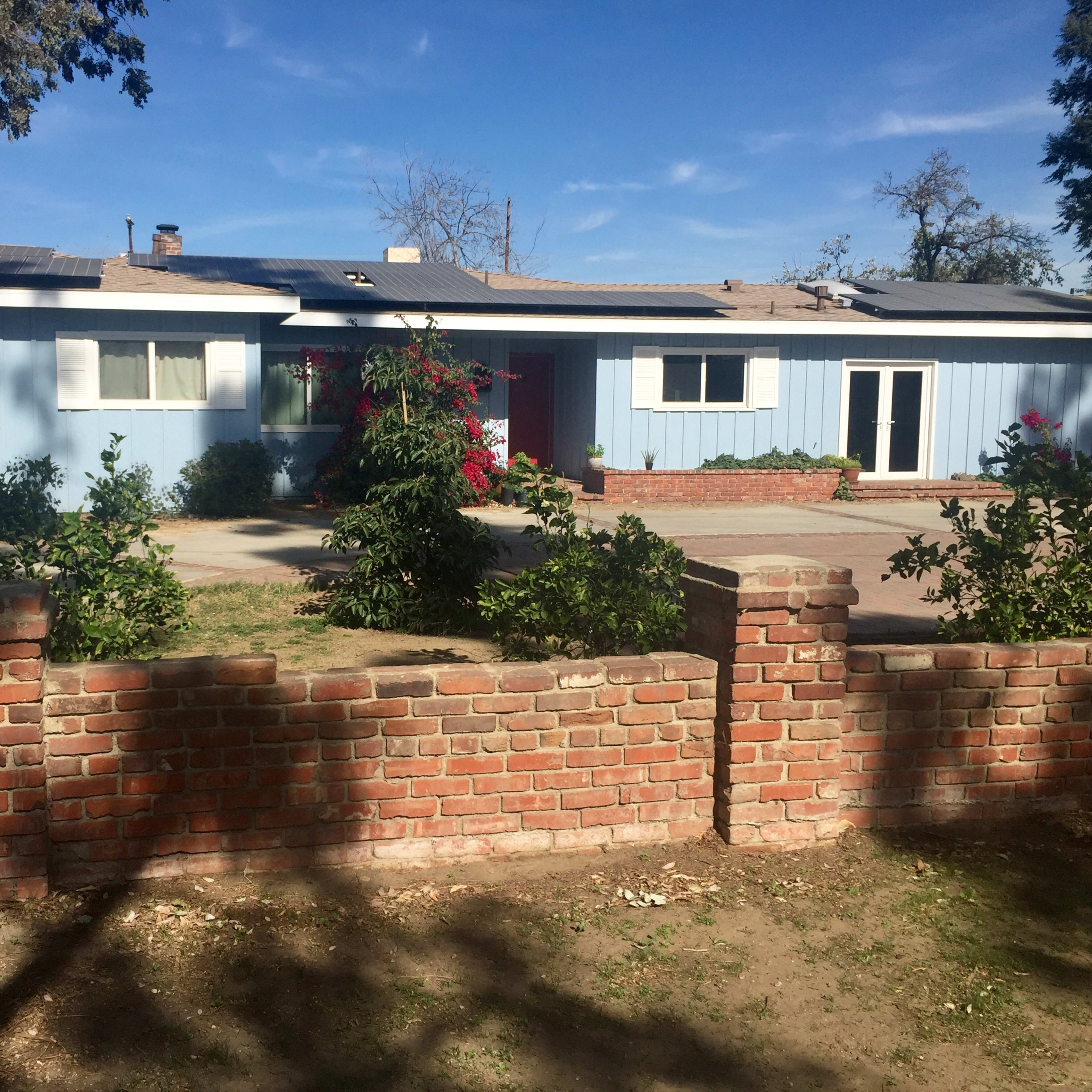 A single-story blue house with a red door is surrounded by a brick wall and landscaped with shrubs and trees.