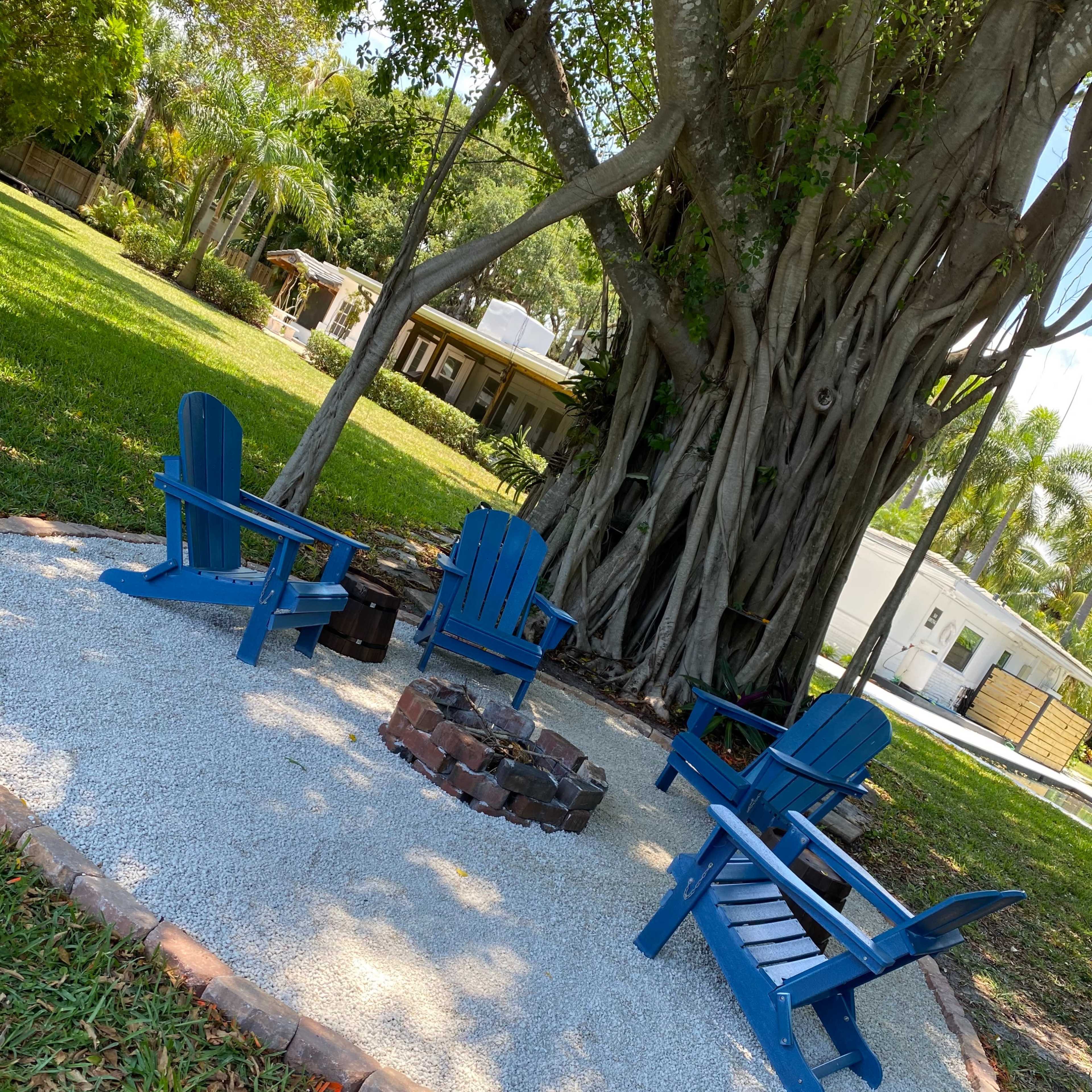 A cozy fire pit area with blue Adirondack chairs is surrounded by gravel and situated under a large tree in a grassy yard.