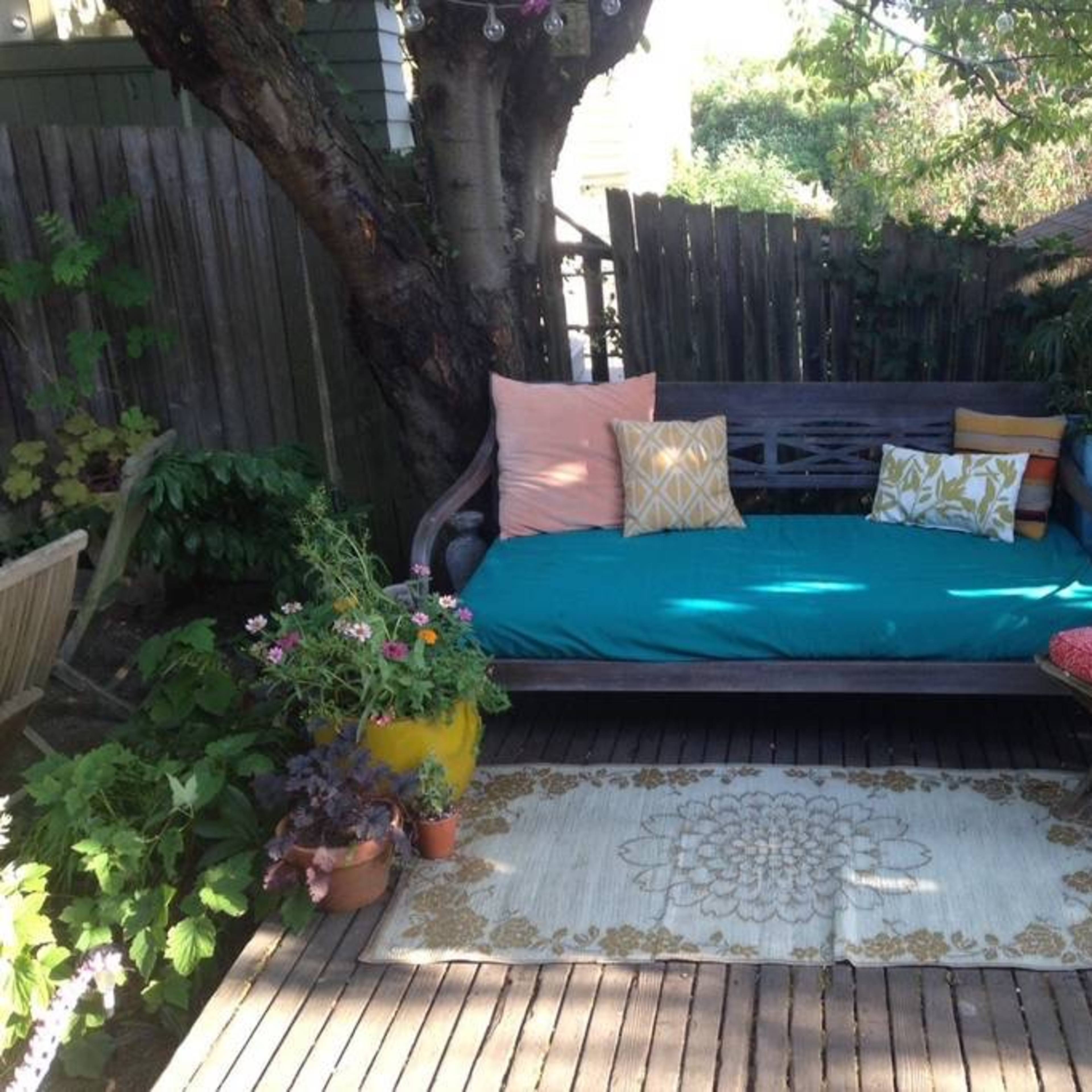 A wooden daybed with colorful pillows, surrounded by potted plants and a decorative rug on a wooden deck.
