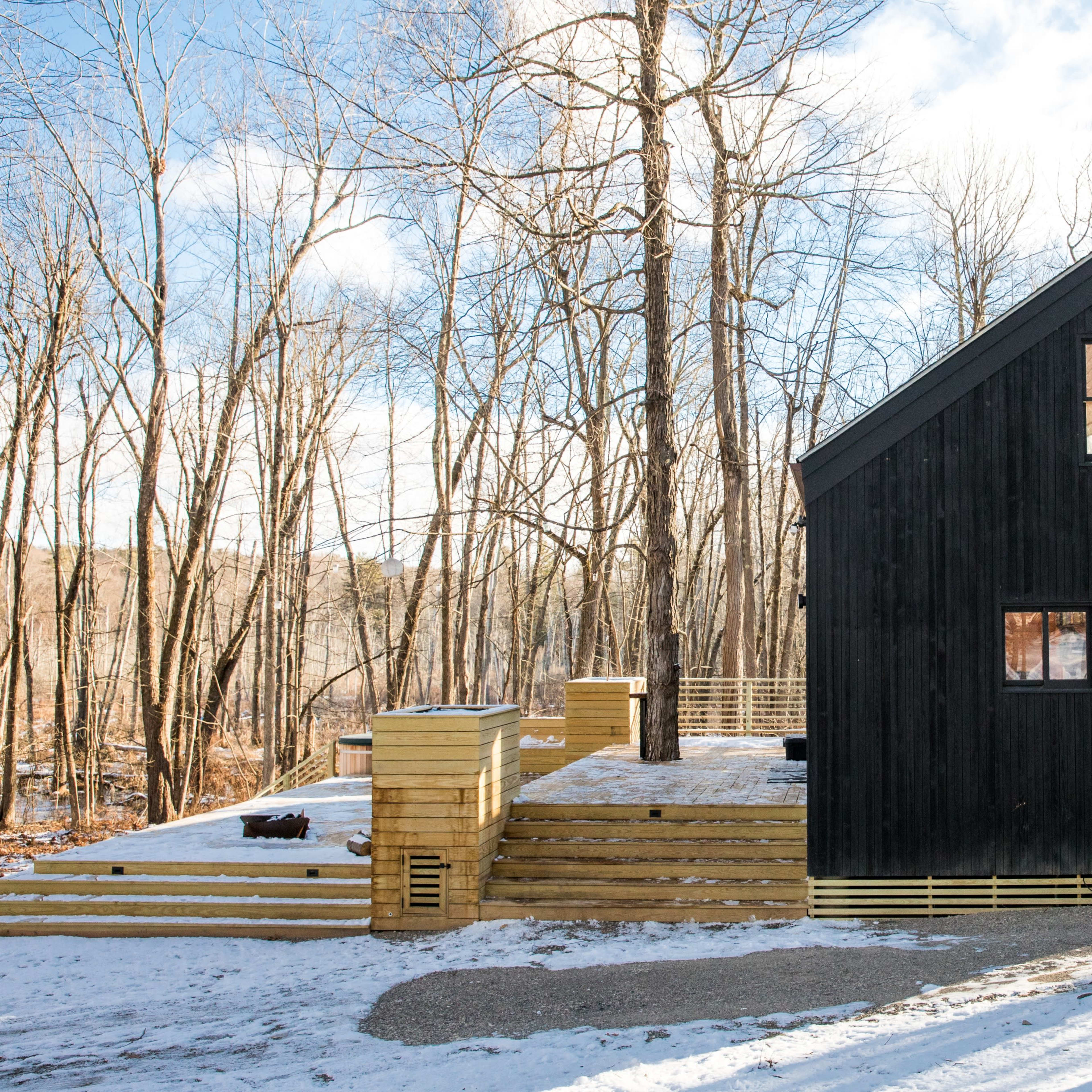 A modern black wooden house with a slanted roof, surrounded by snow and trees in a wooded area.