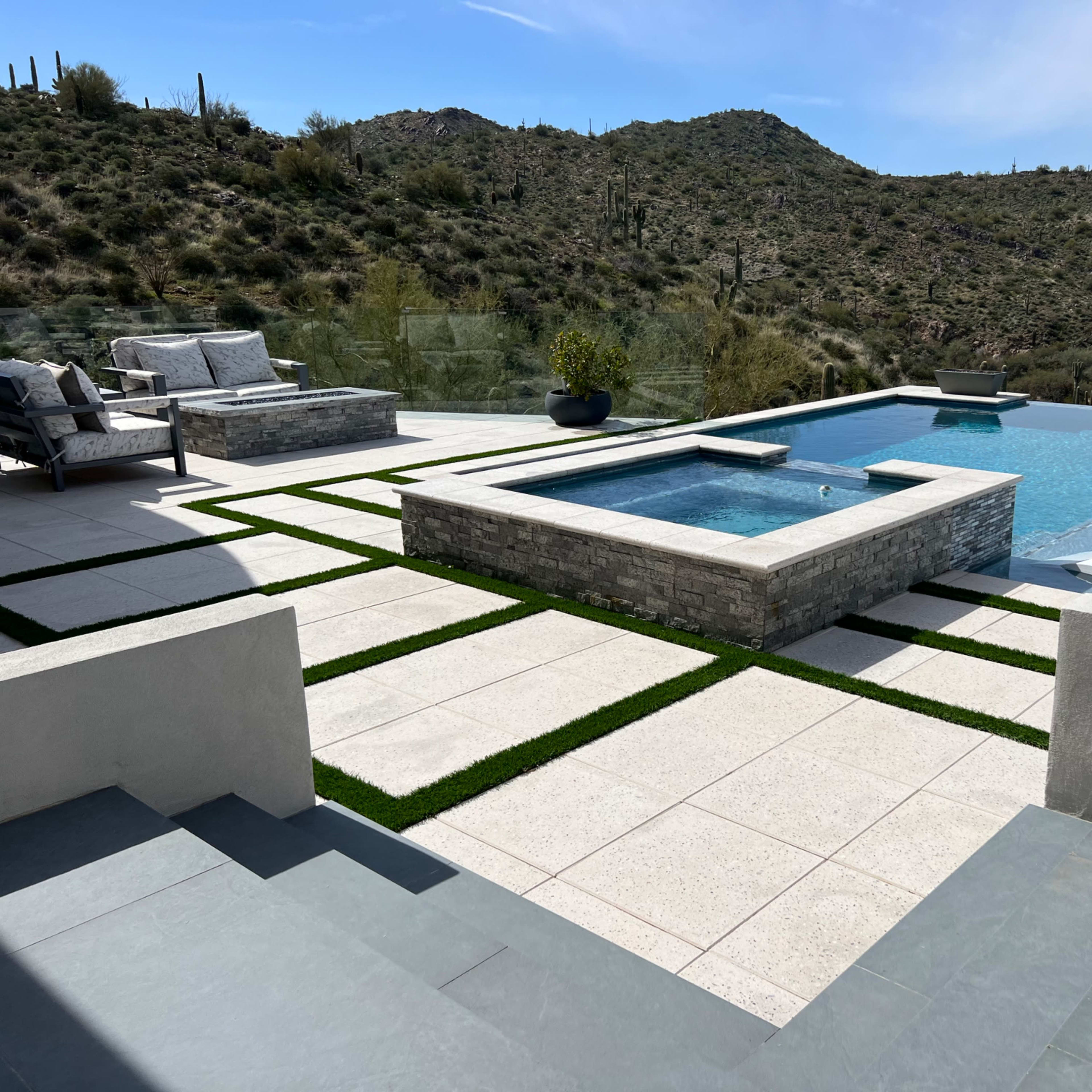 A modern outdoor pool area with a water feature, lounge chairs, and a view of cacti-covered hills in the background.