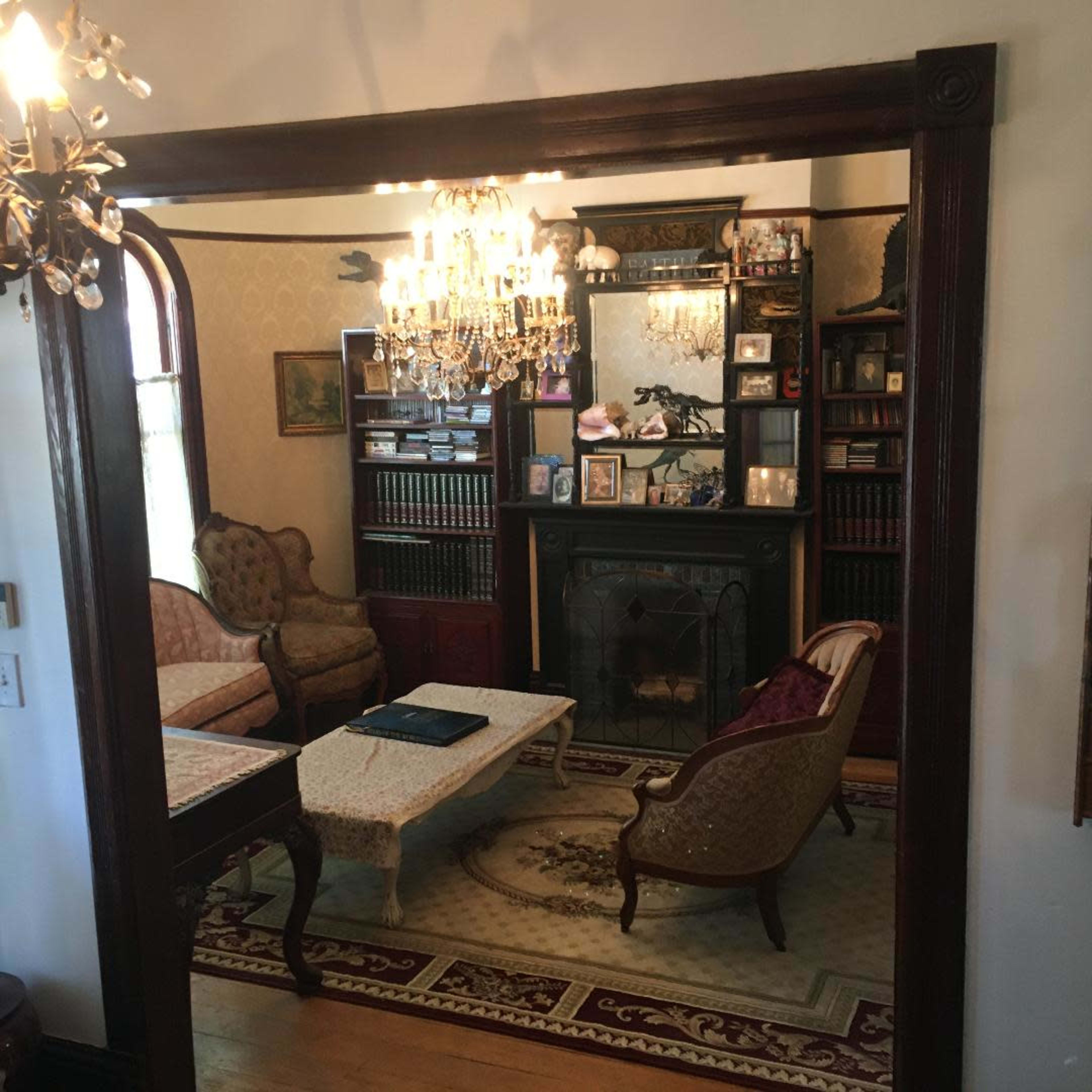 A living room with antique furniture, including a couch and armchairs, centered around a coffee table, and features a fireplace and bookshelves along the back wall.