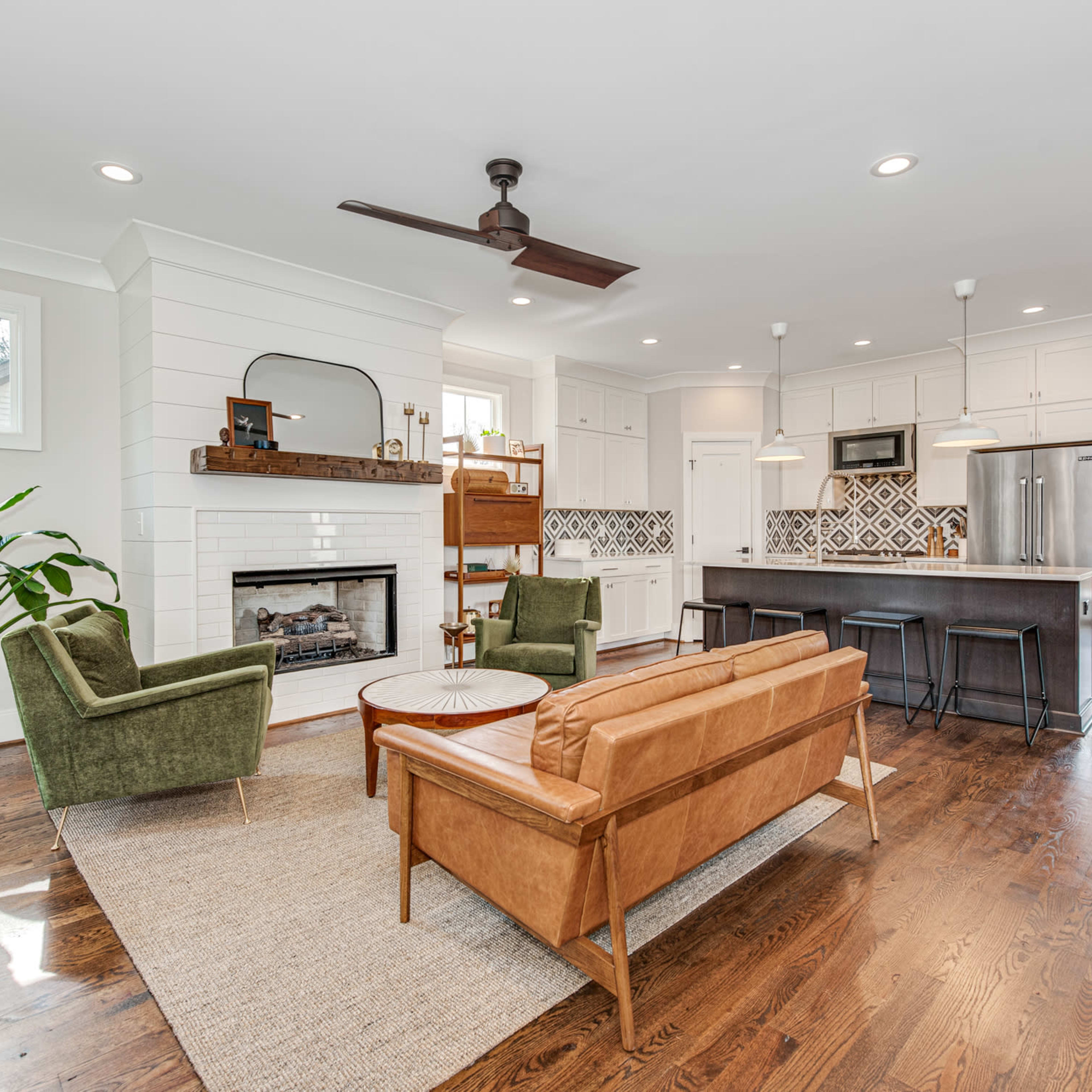 A modern living room features a brown leather sofa, green armchairs, a fireplace, and a kitchen area with patterned tiles and bar stools.
