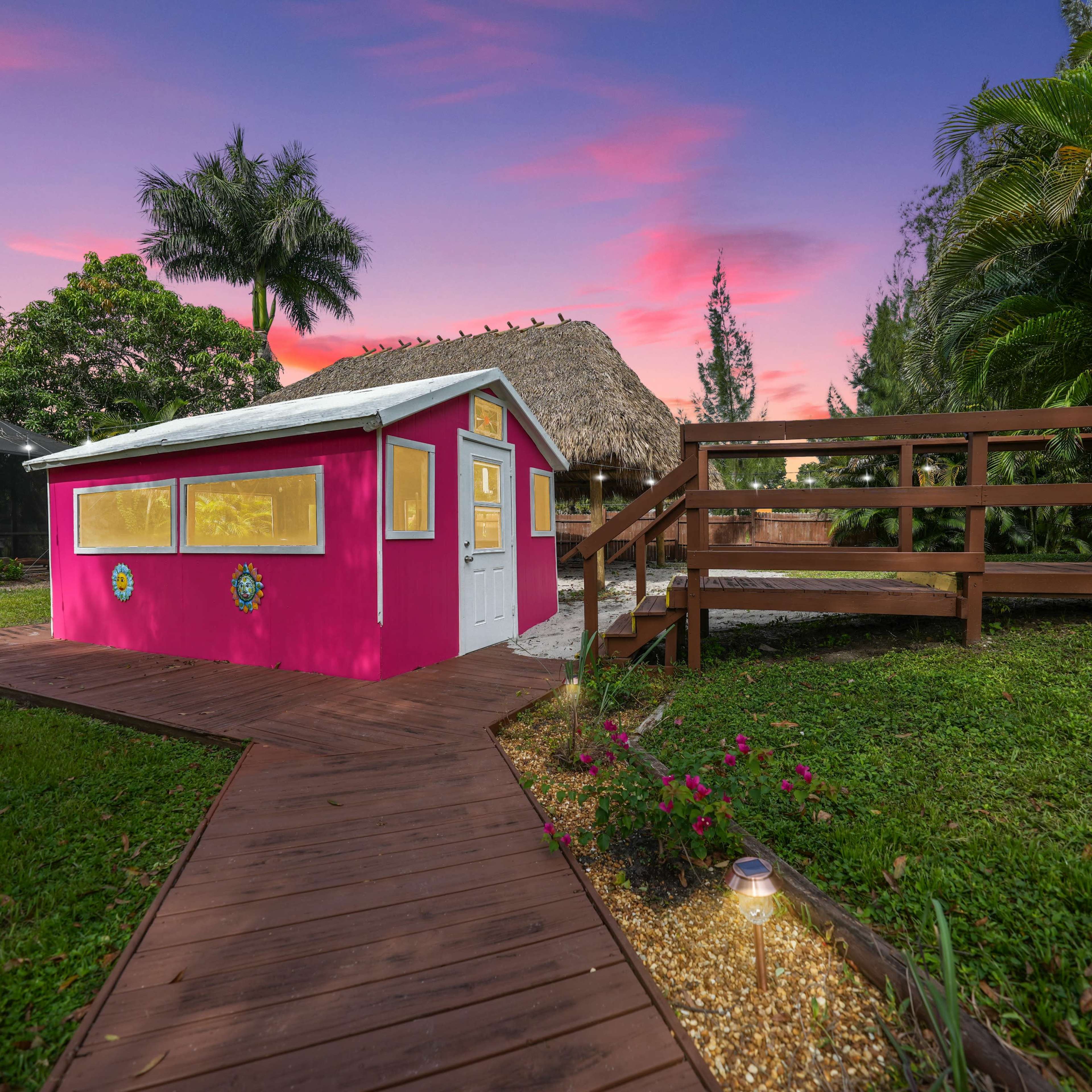 A colorful pink shed with yellow windows situated next to a thatched-roof structure, surrounded by lush greenery and a wooden walkway leading to a raised deck area at sunset.