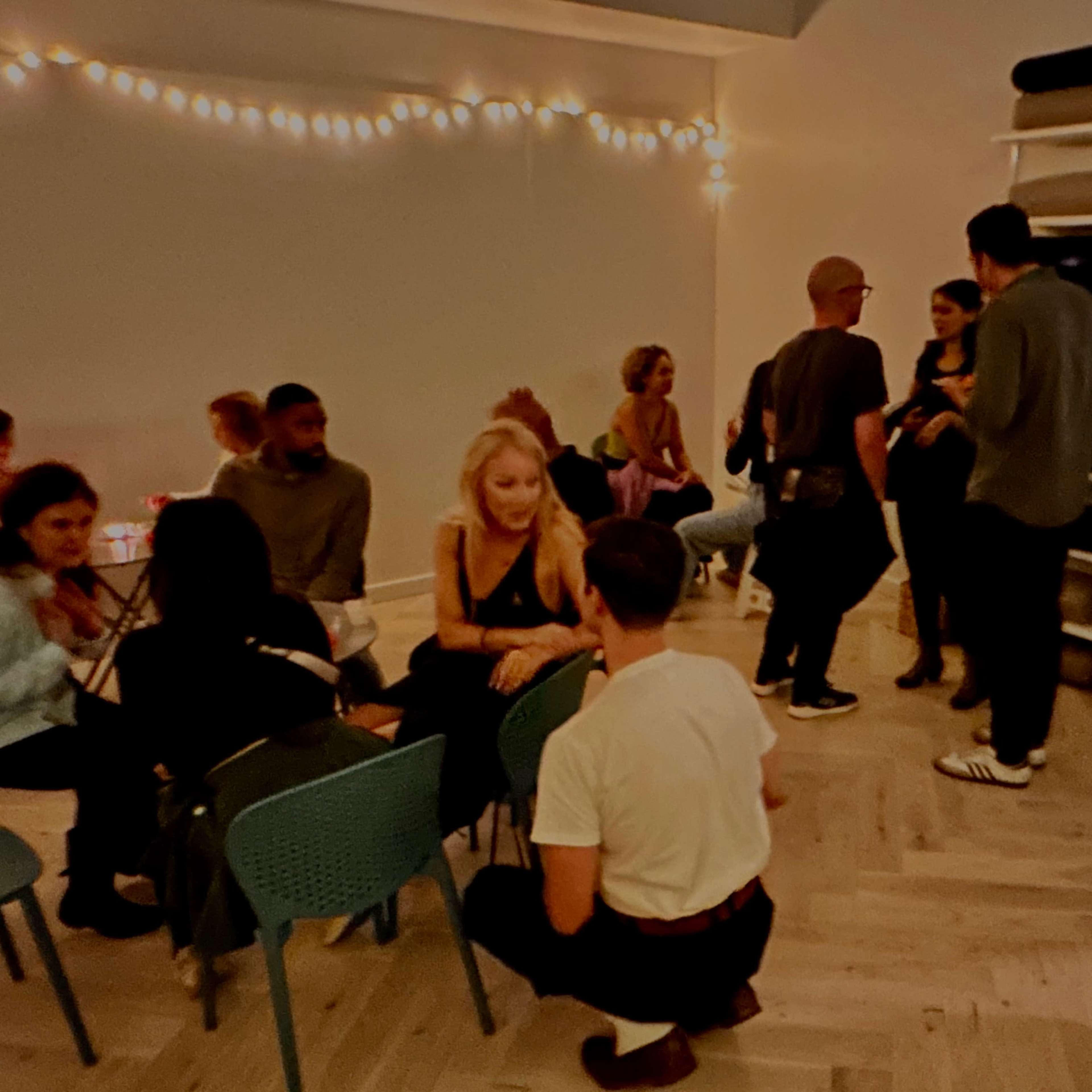 A group of people socializes in a well-lit room decorated with string lights, seated at tables and standing in conversation.