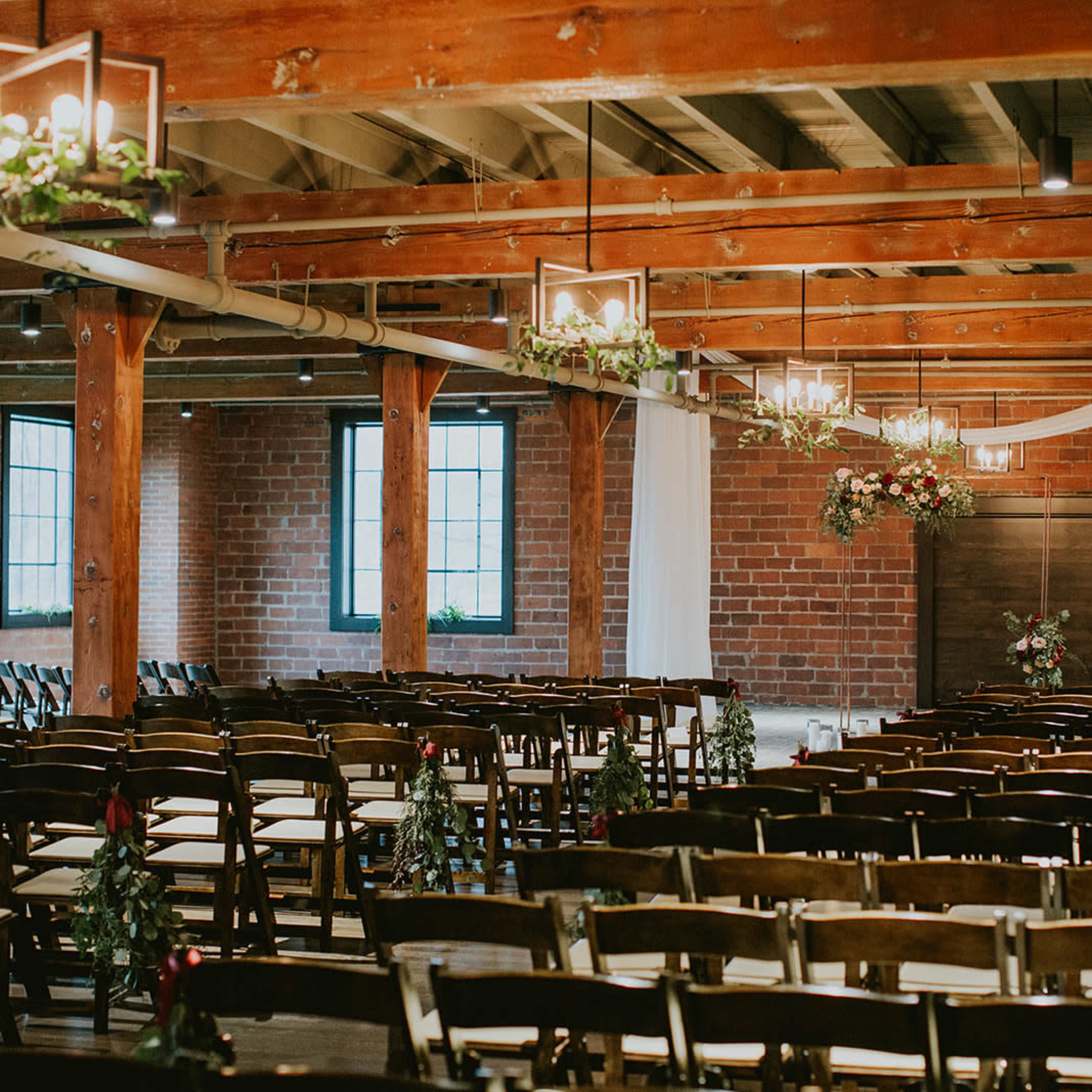 A wedding venue with rows of wooden chairs arranged facing a decorative backdrop and exposed brick walls, featuring hanging lights and greenery.