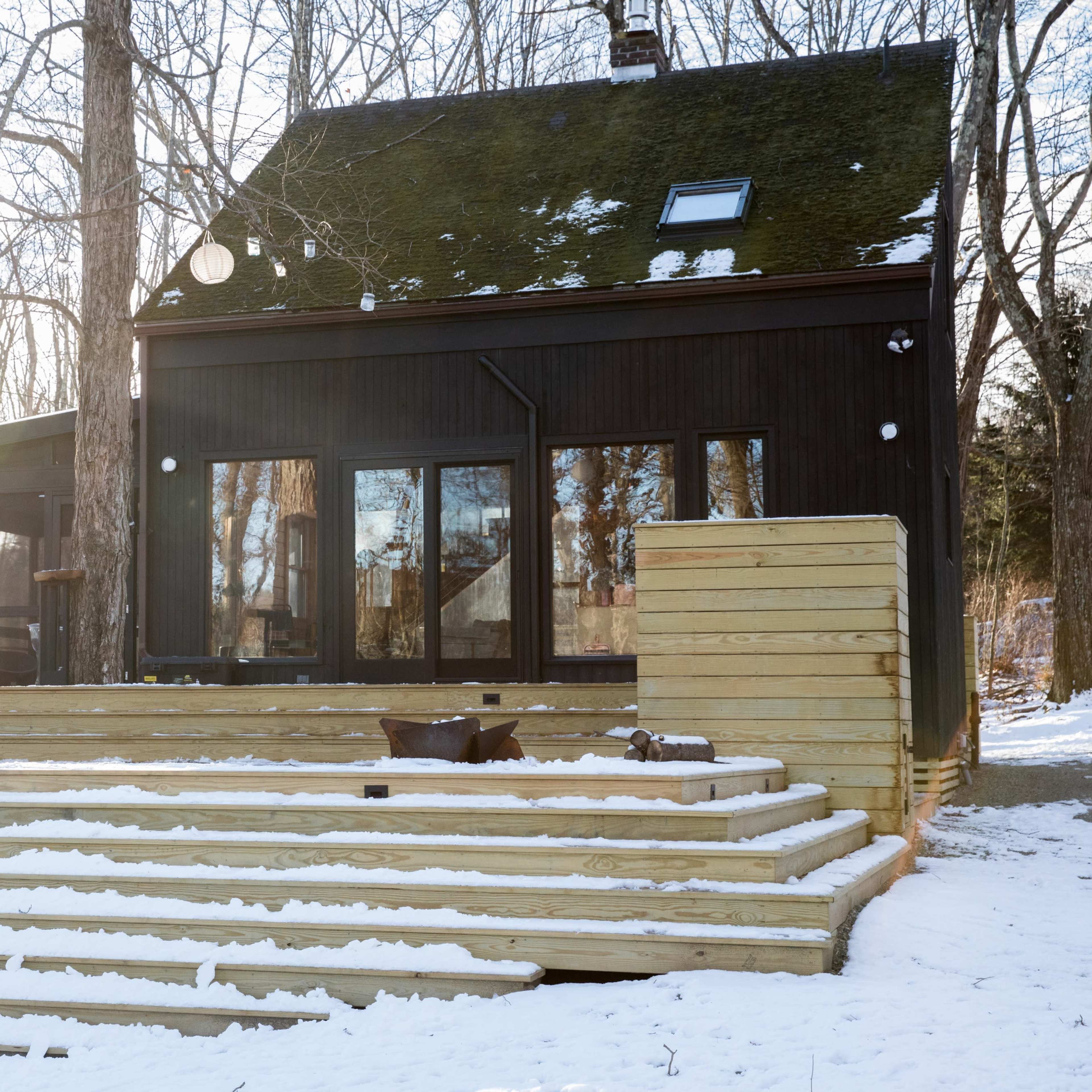 A contemporary wooden house with large windows and a sloped roof is situated on a snow-covered deck surrounded by bare trees.