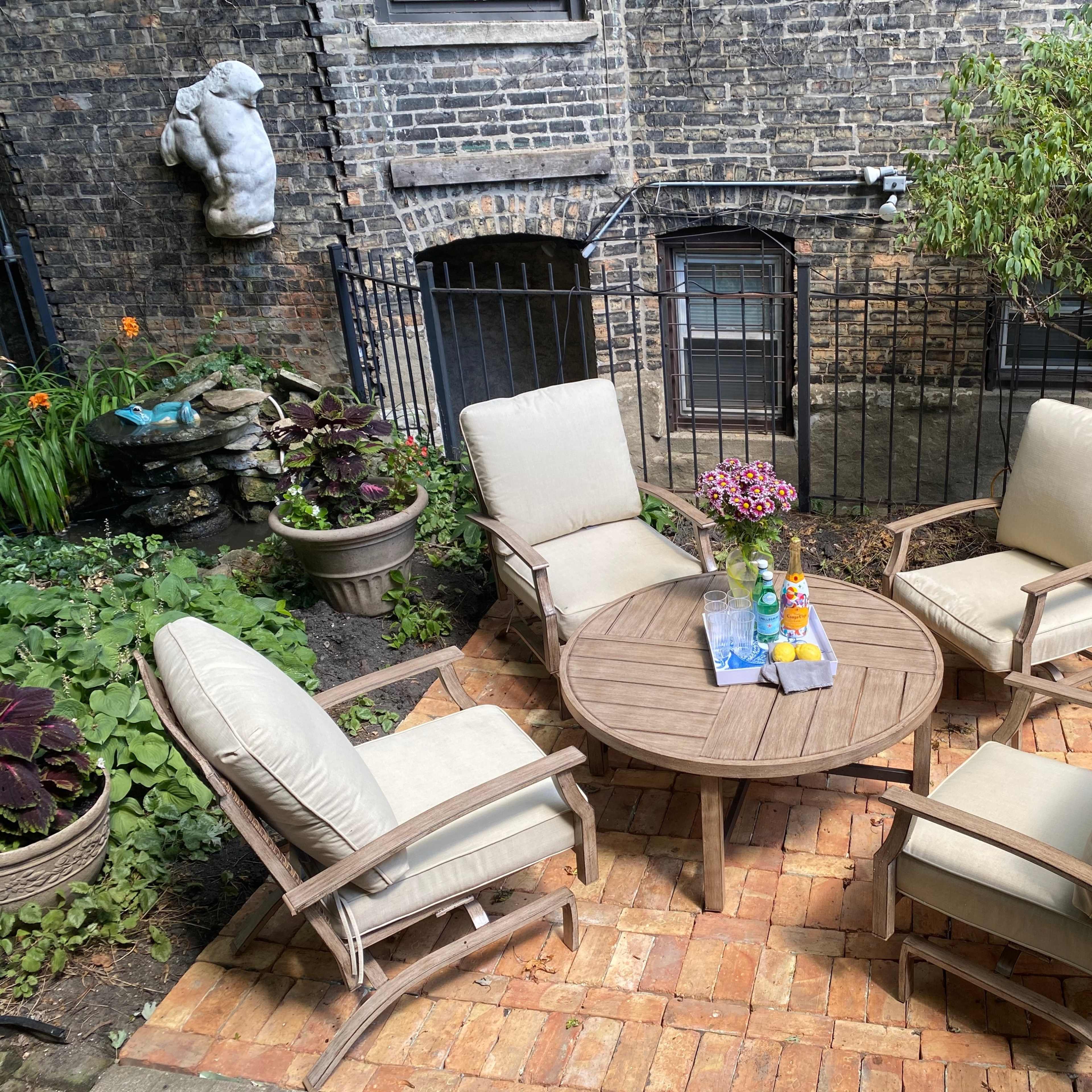 A circular wooden table surrounded by four cushioned chairs sits in a brick-paved outdoor space, with plants and a stone feature in the background.