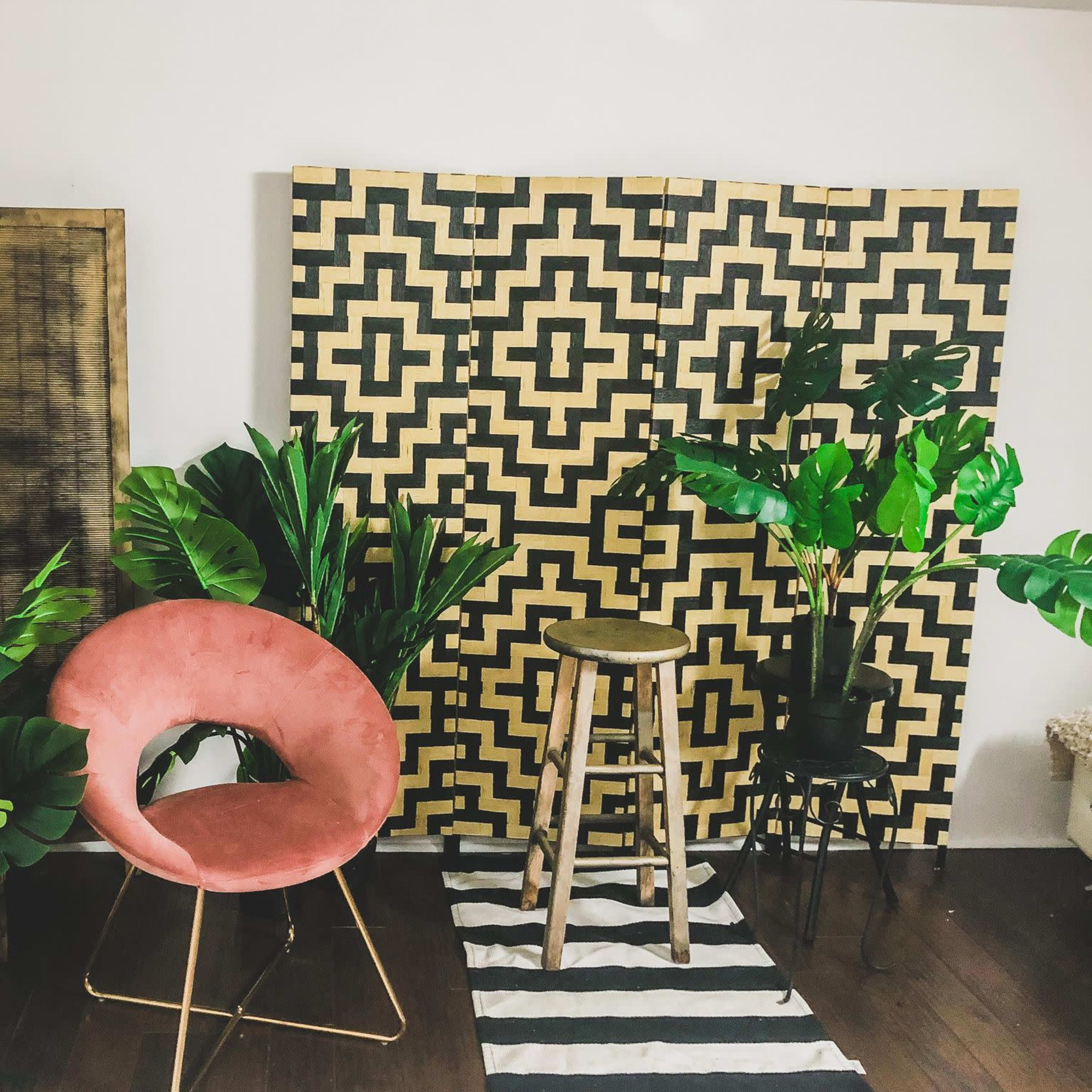 A modern room featuring a pink chair, a wooden stool, and various green plants against a patterned backdrop.