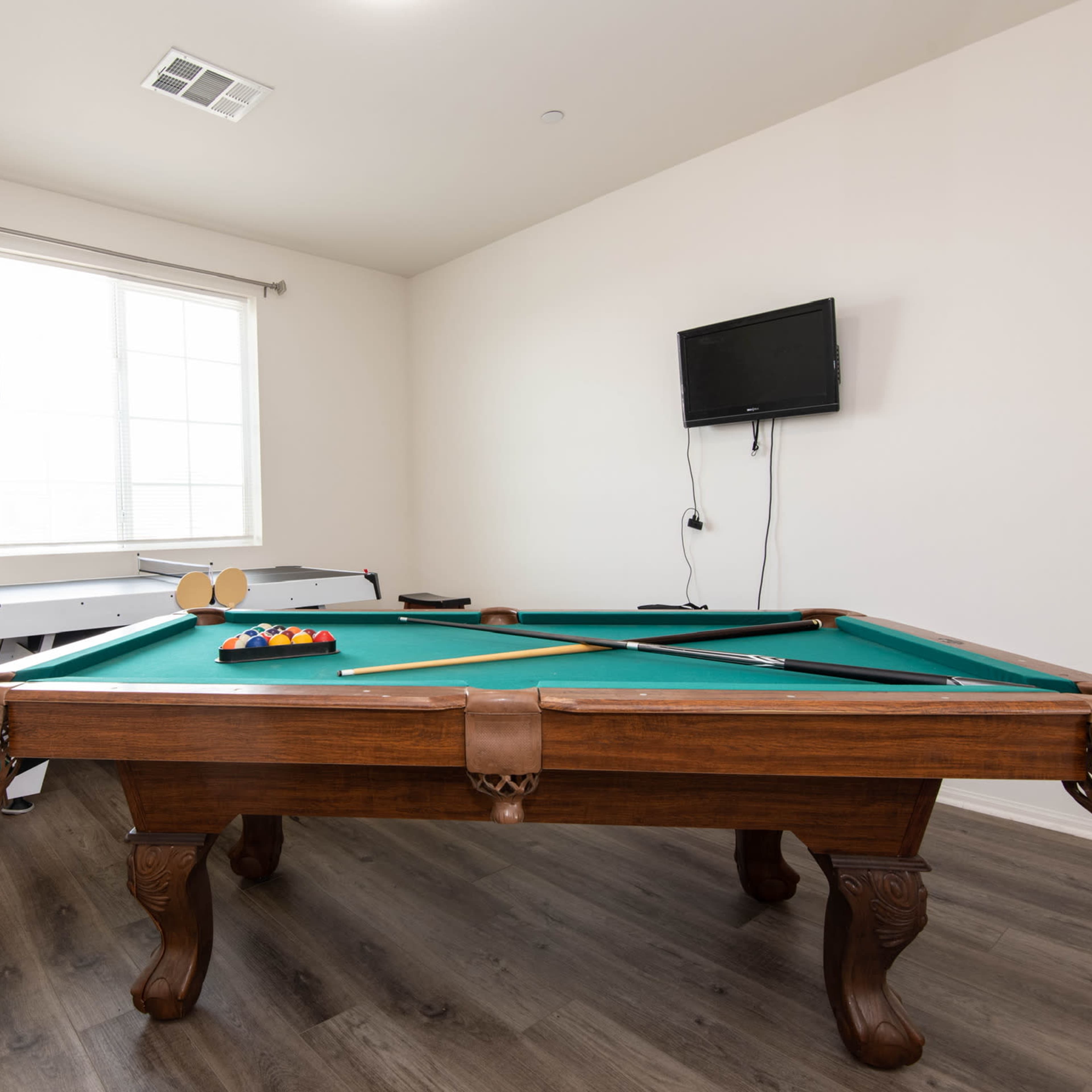 A billiards table and an air hockey table are positioned in a well-lit room with a window and a mounted television.