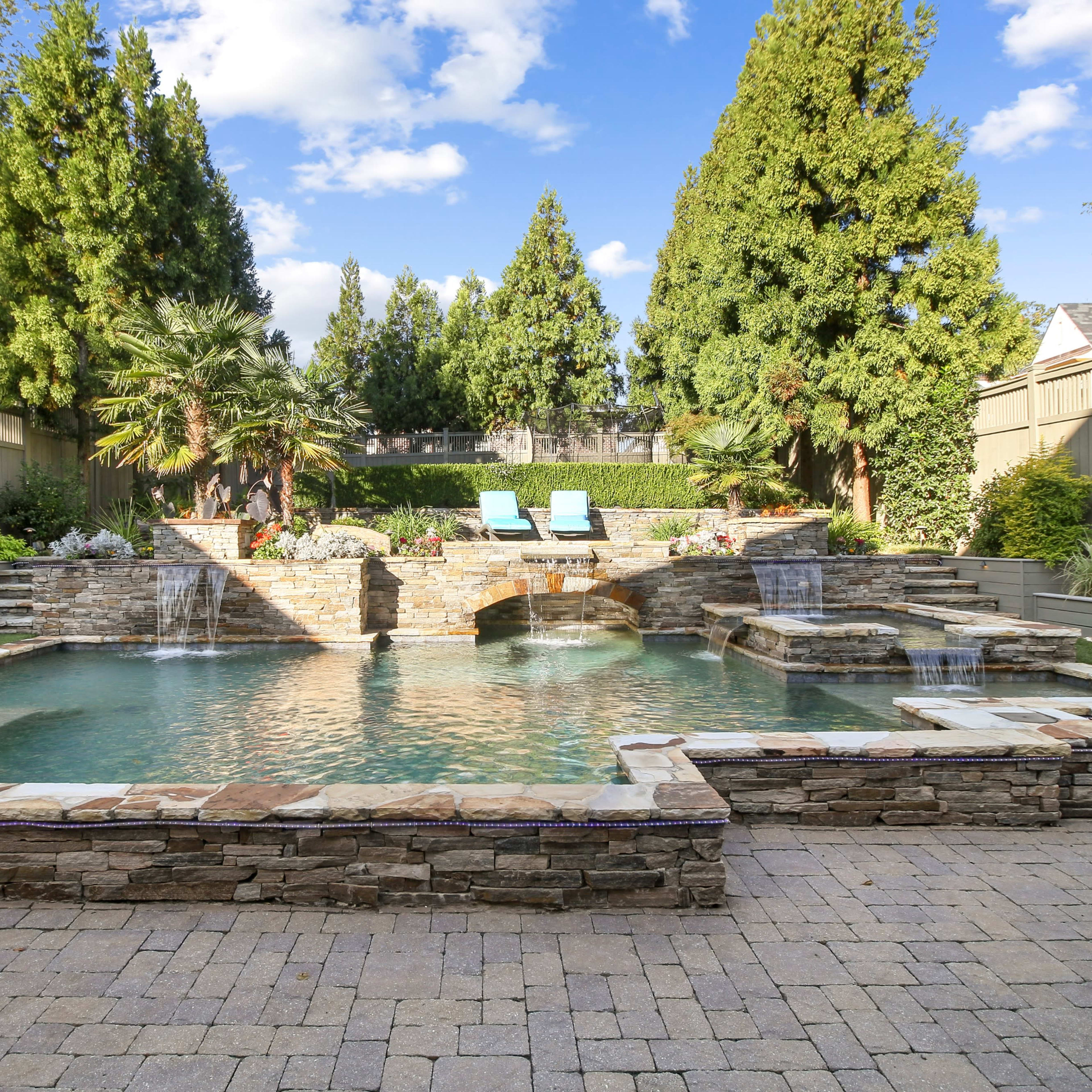 A landscaped backyard featuring a stone-accented swimming pool with waterfalls, surrounded by lush greenery and lounge chairs.