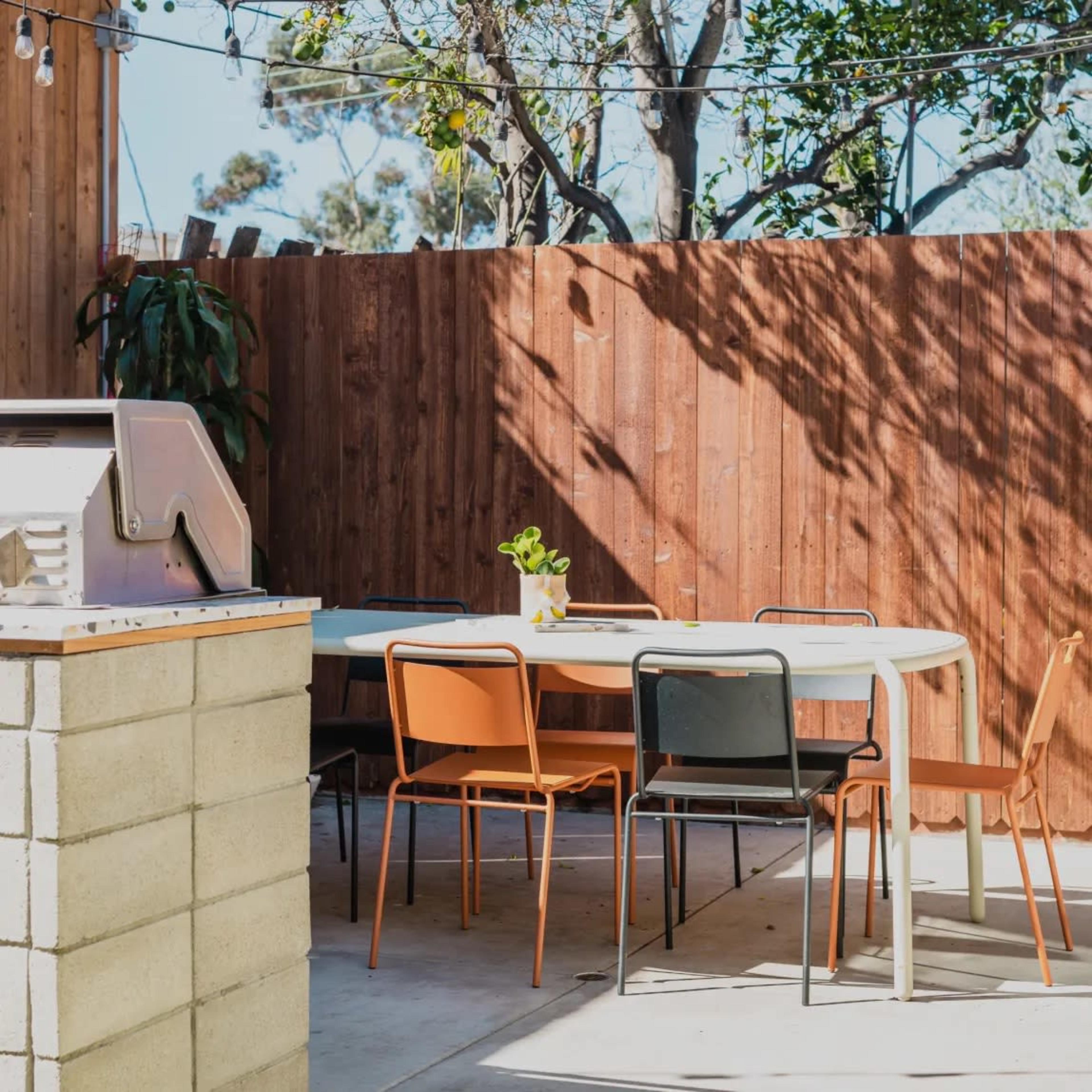 A patio area features a table surrounded by colorful chairs, with a grill and wooden fencing in the background.
