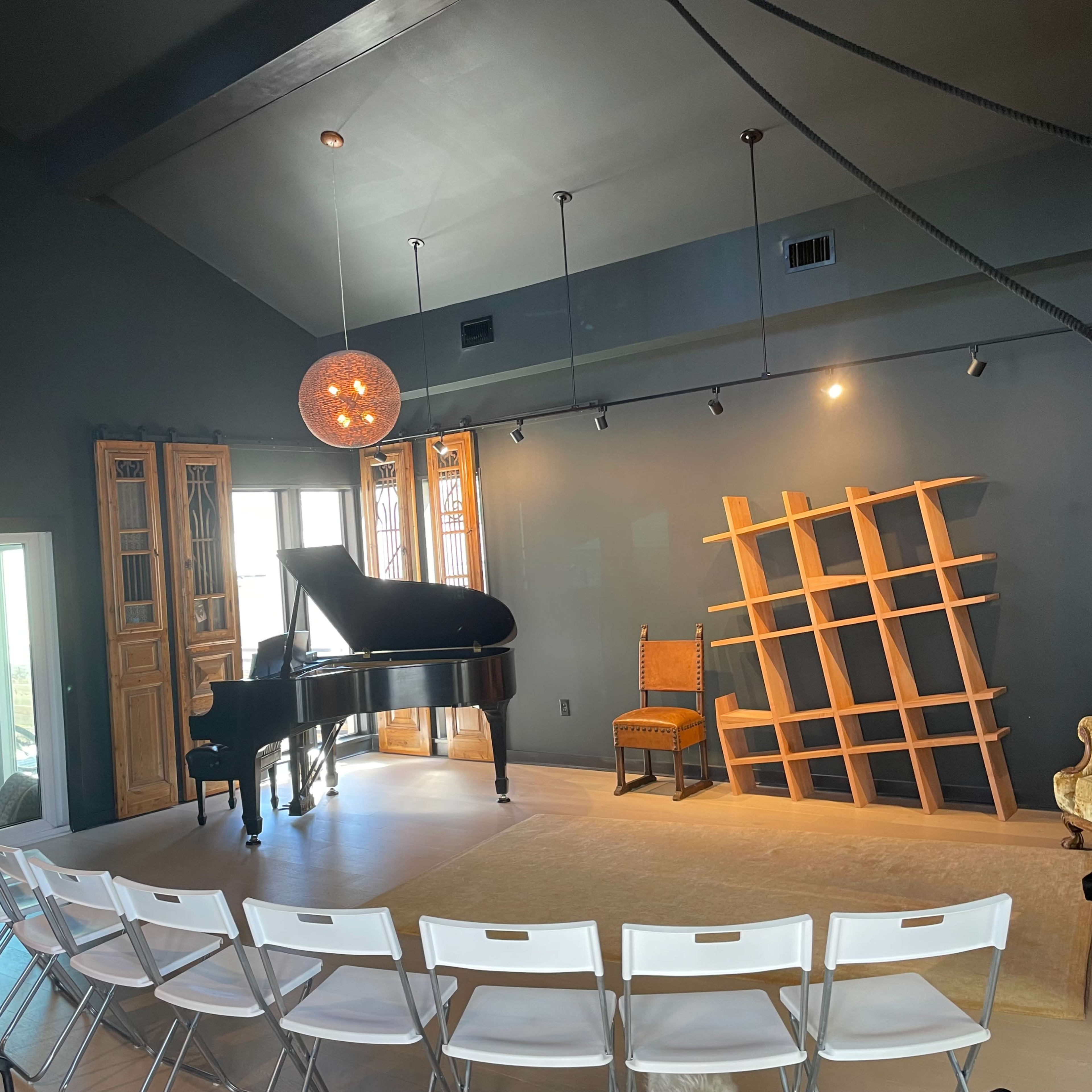 A black piano is positioned in the center of a room with gray walls, accompanied by a wooden chair and a shelving unit against the wall, while white folding chairs are set up in front.