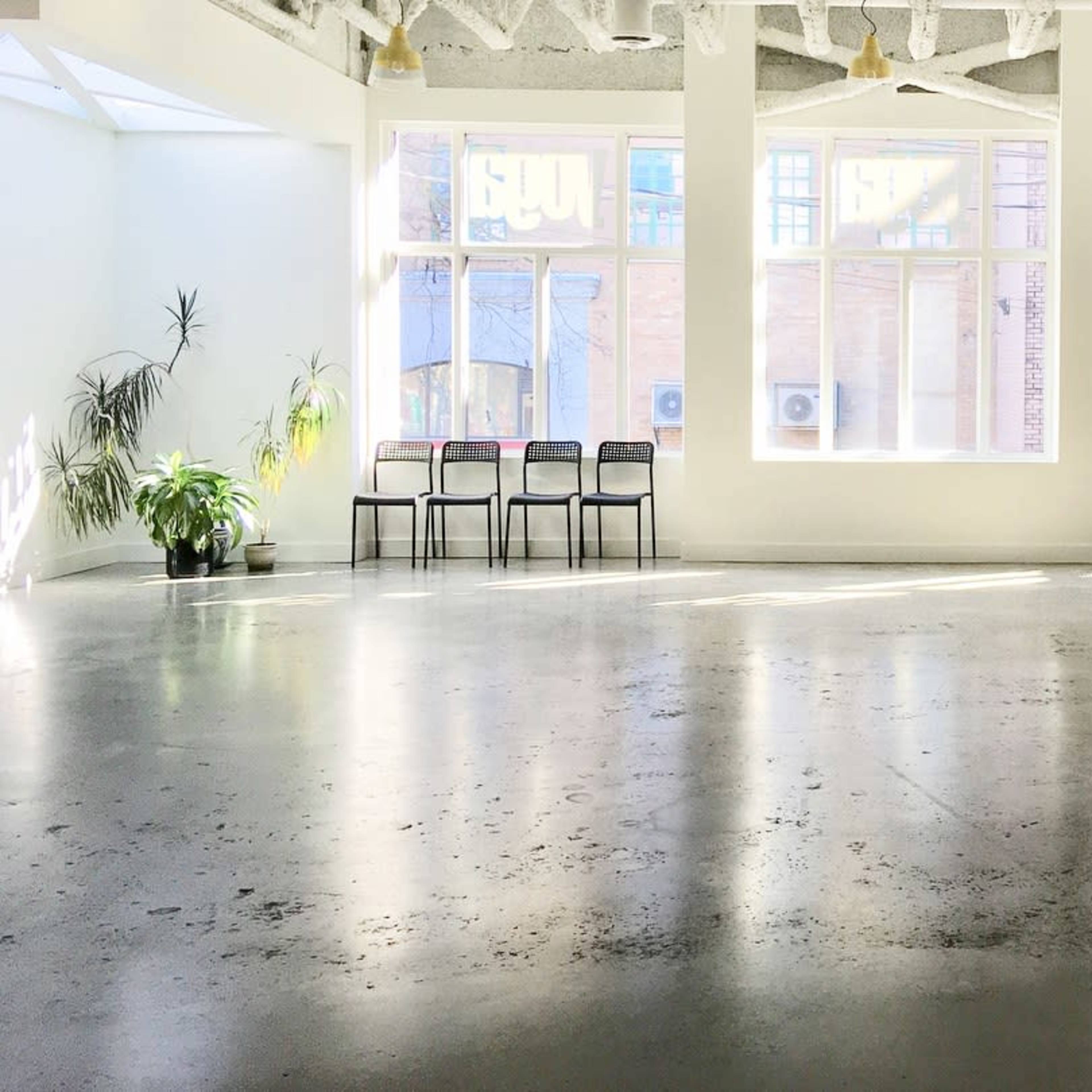 An empty room with concrete floors, several large windows allowing natural light, and a row of black chairs along one wall.