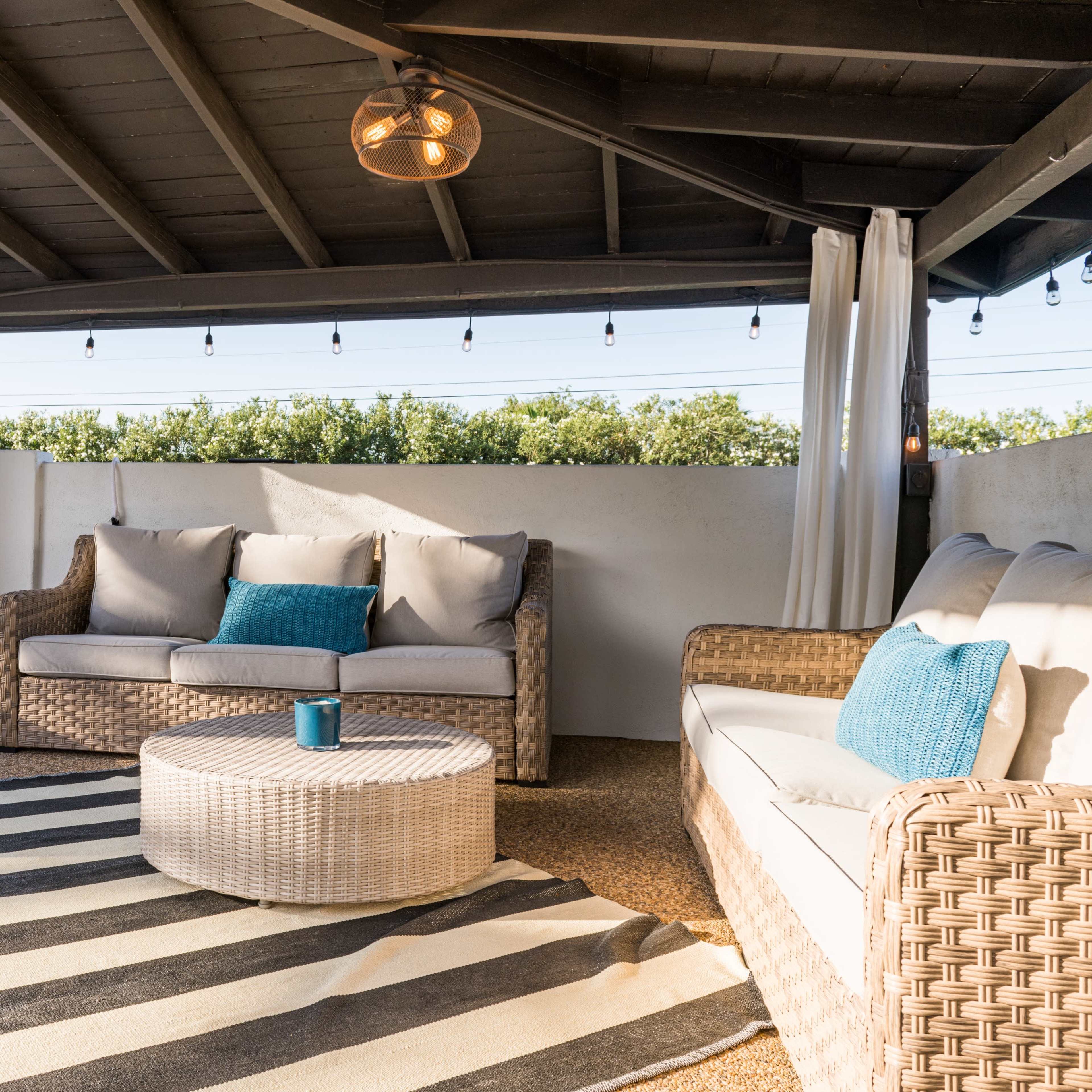 A covered outdoor seating area features a sofa and lounge chair on a patterned rug, with a round table in the center.