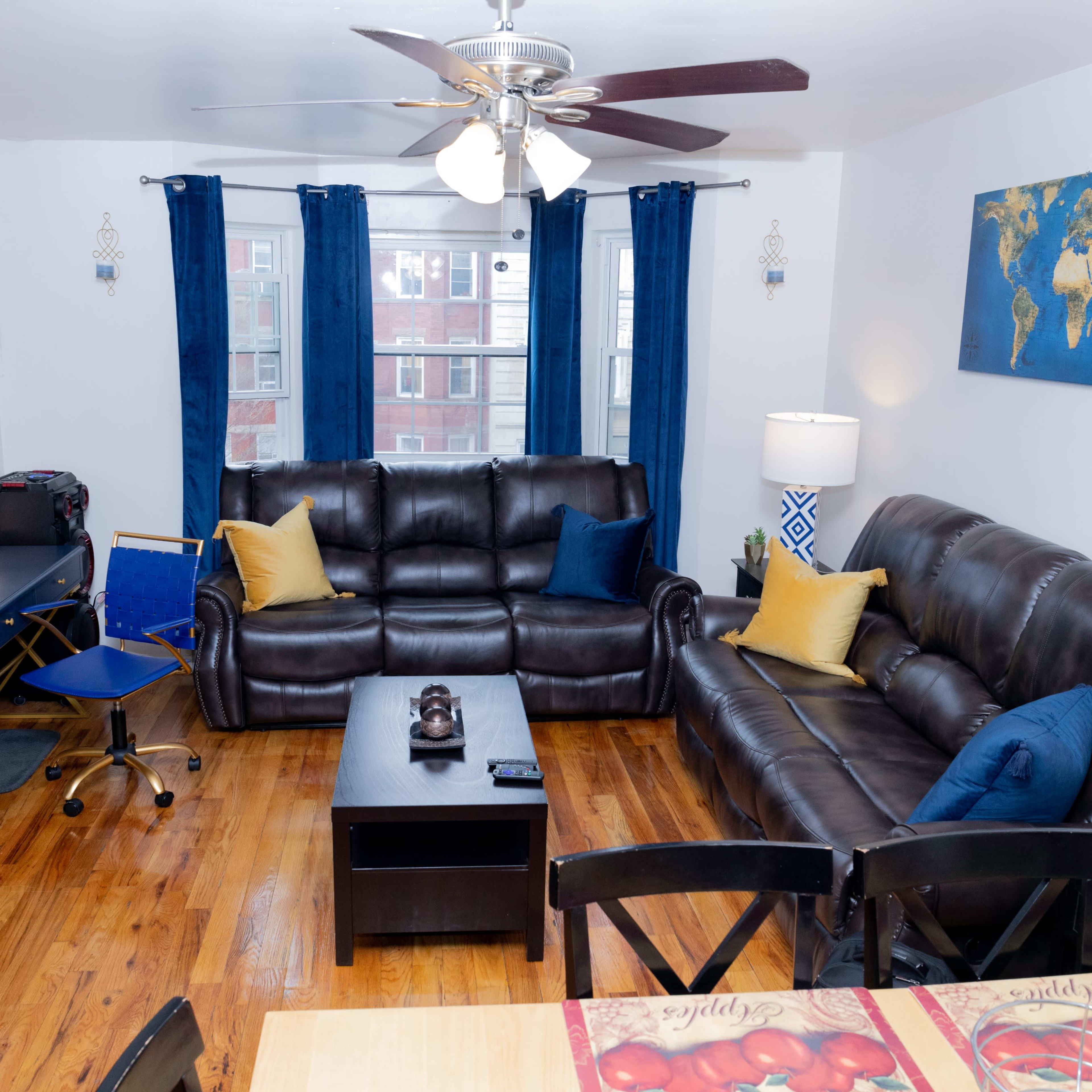 A living room features two dark leather sofas, a coffee table, a TV stand, and a desk, with blue curtains framing the windows and a world map on the wall.