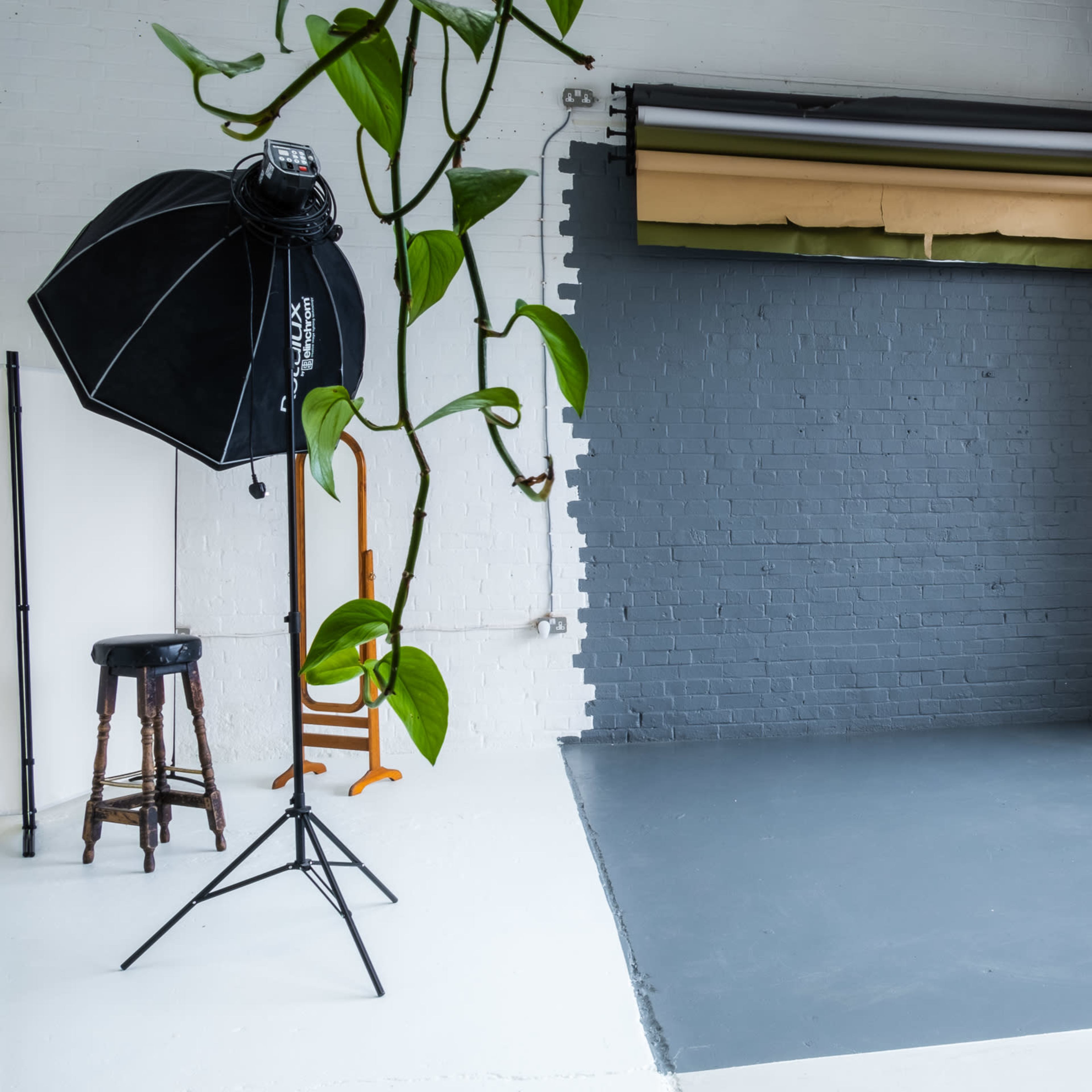 A photography studio with a gray wall, a light stand, a tall stool, and a window letting in natural light.