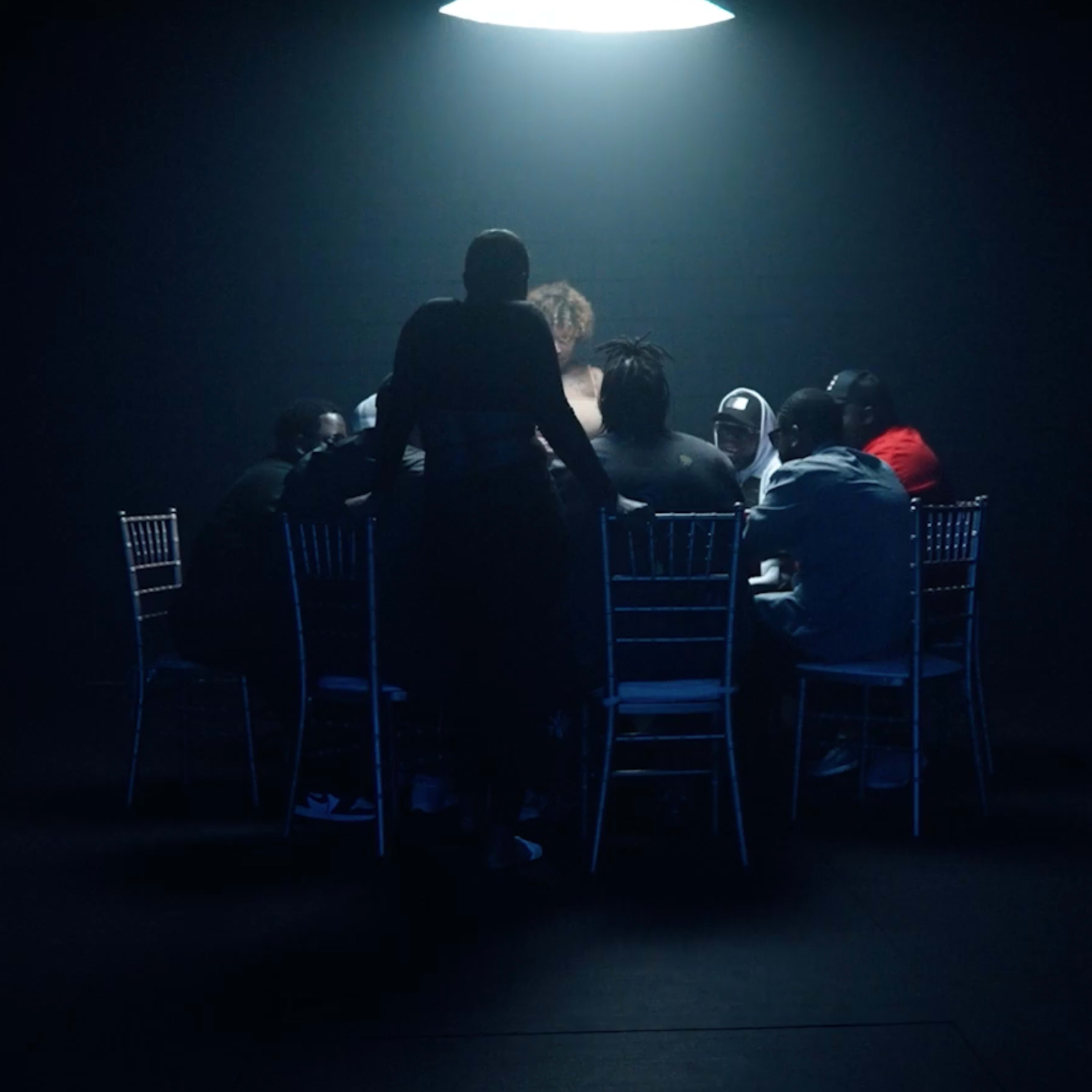 In a dimly lit room, a group of seated individuals surrounds a table under a single overhead light.