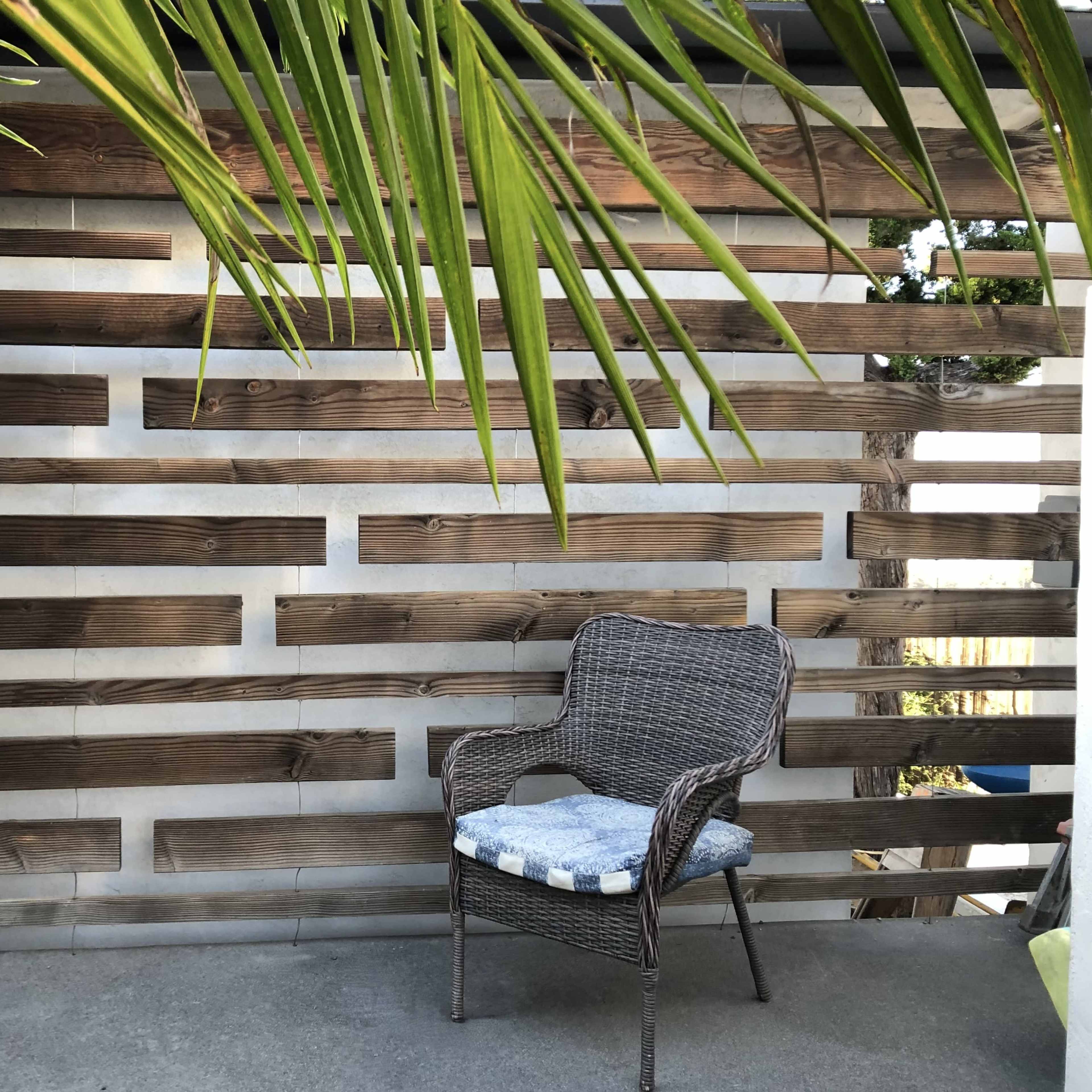 A wicker chair sits against a wooden slat wall, flanked by green palm leaves.