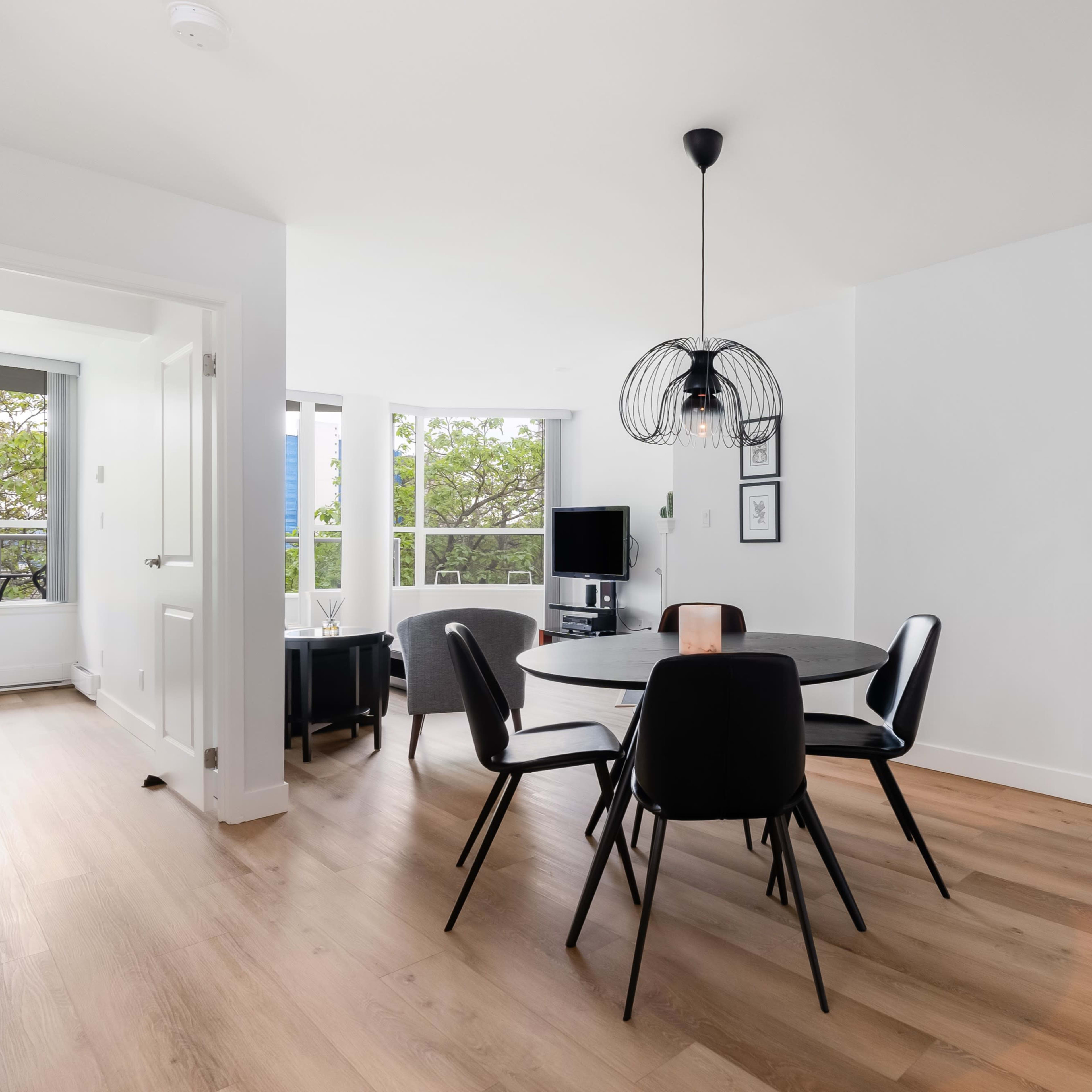 A modern dining area features a round table surrounded by black chairs, with large windows allowing natural light and greenery to view outside.