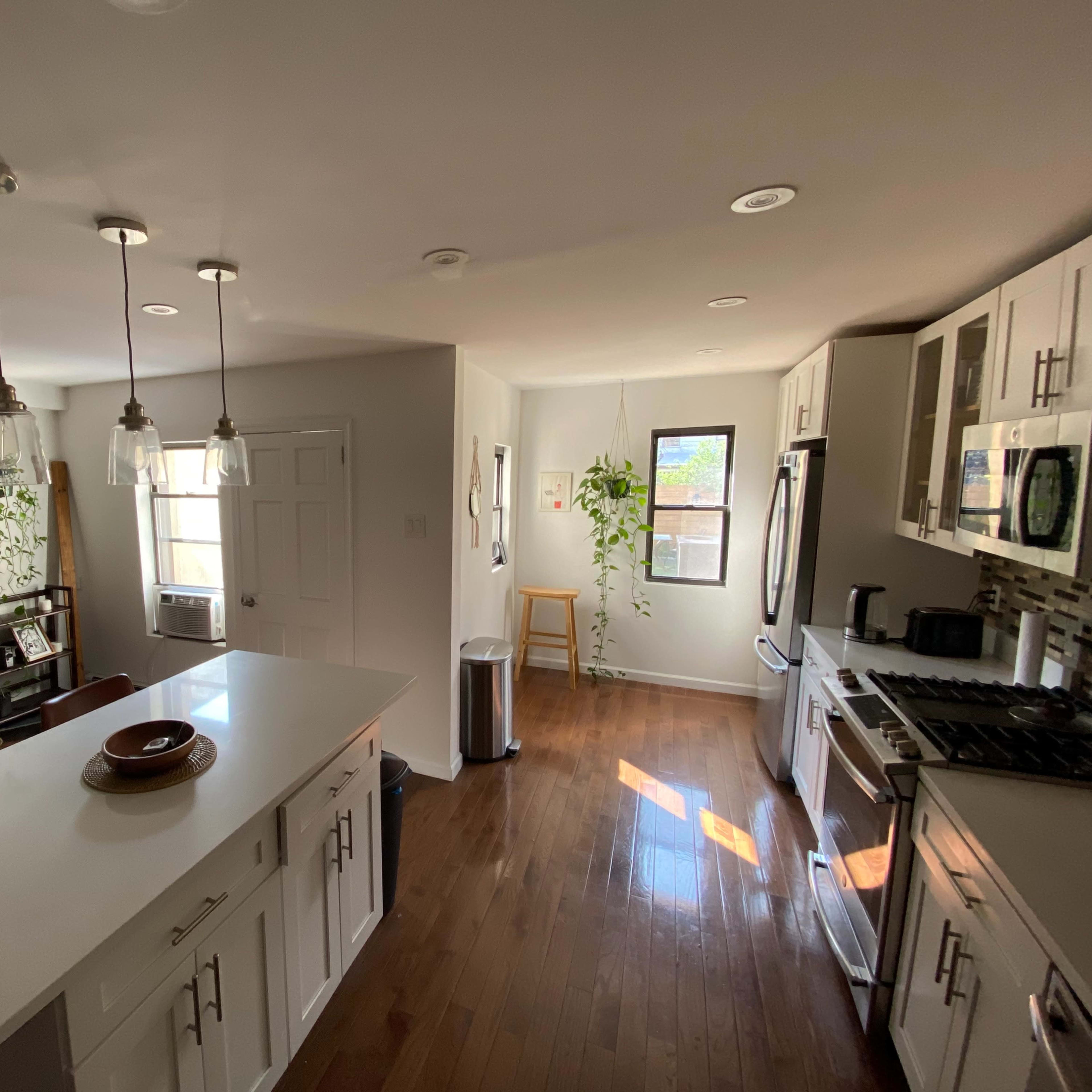 A modern kitchen with a central island, equipped with a stove, refrigerator, and an eating area, alongside an adjacent living space with a chair and a window.