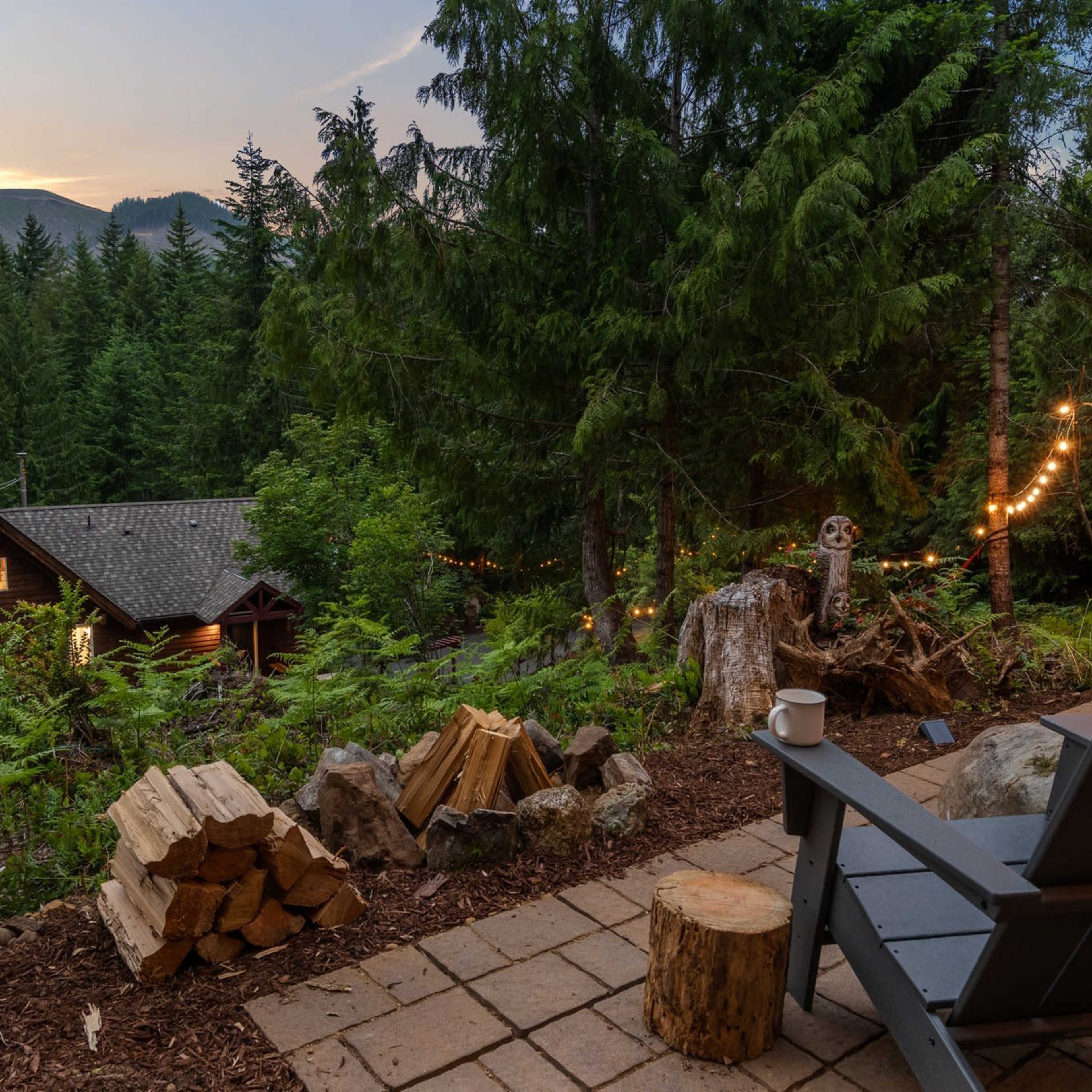 A cozy outdoor seating area with an Adirondack chair overlooking a forested landscape and a cabin, illuminated by string lights.