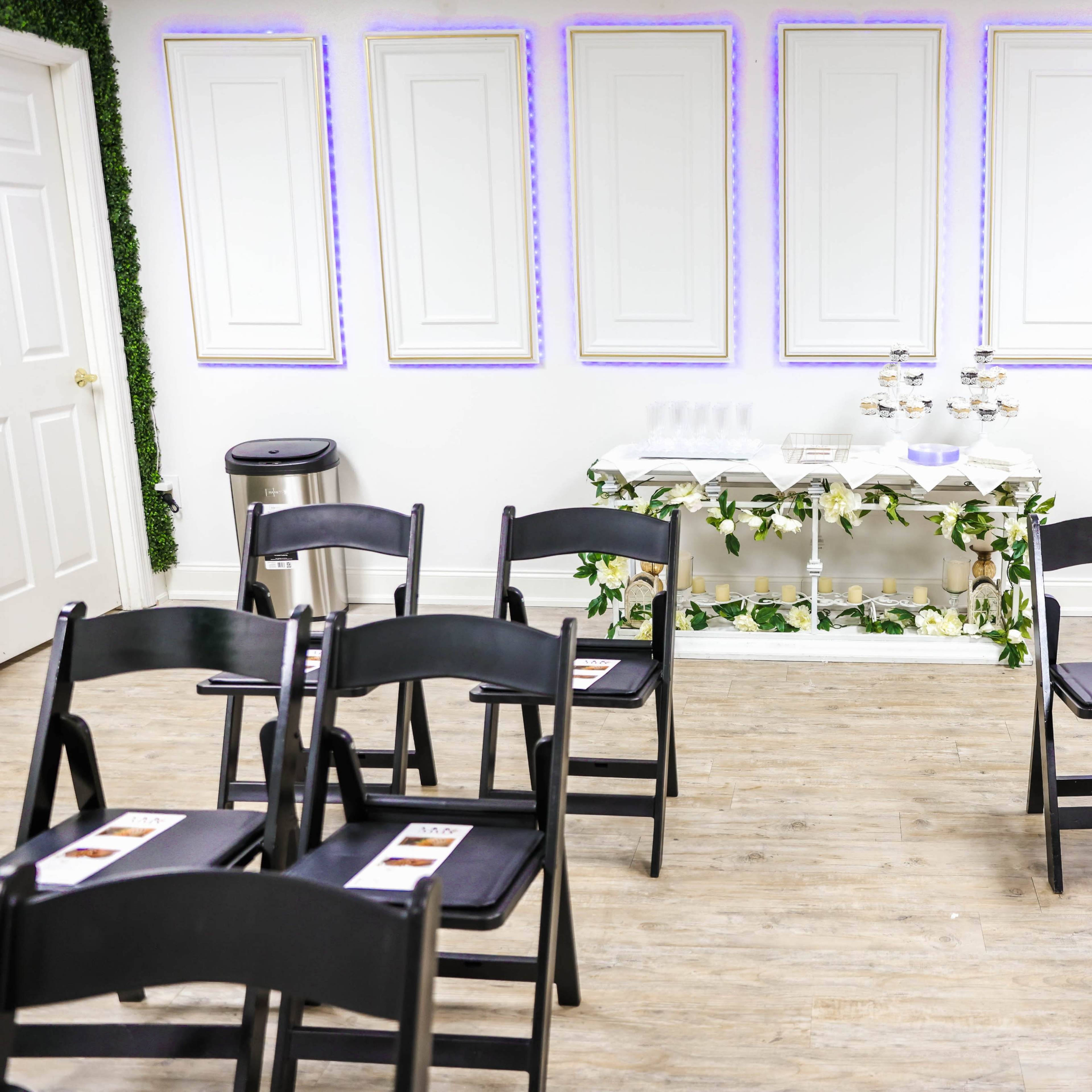 A room set up for an event, with several black chairs arranged in rows facing a decorated table against a white wall.