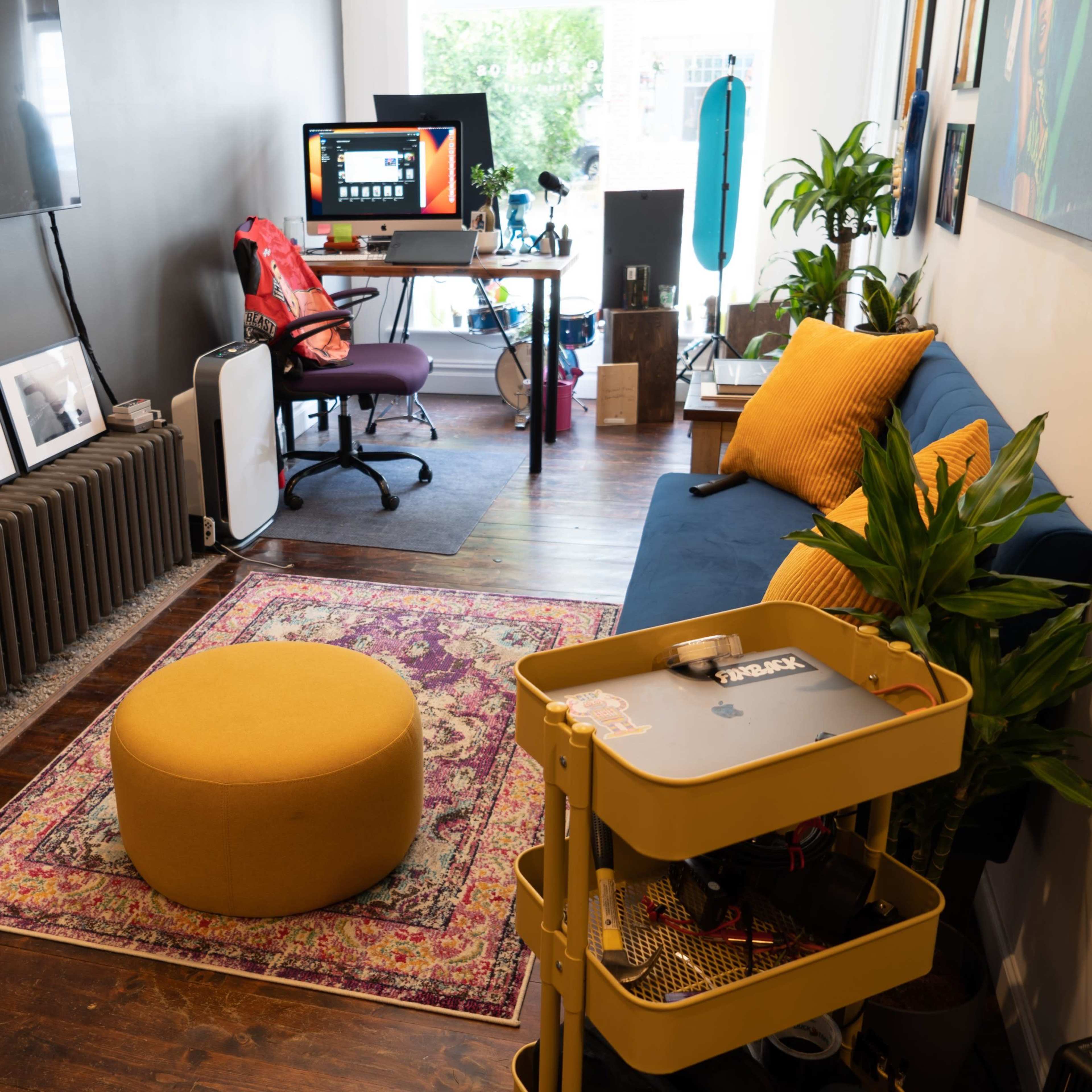 A cozy room featuring a blue sofa, a yellow ottoman, several framed photographs on the wall, a desk with a computer setup, and plants placed throughout the space.