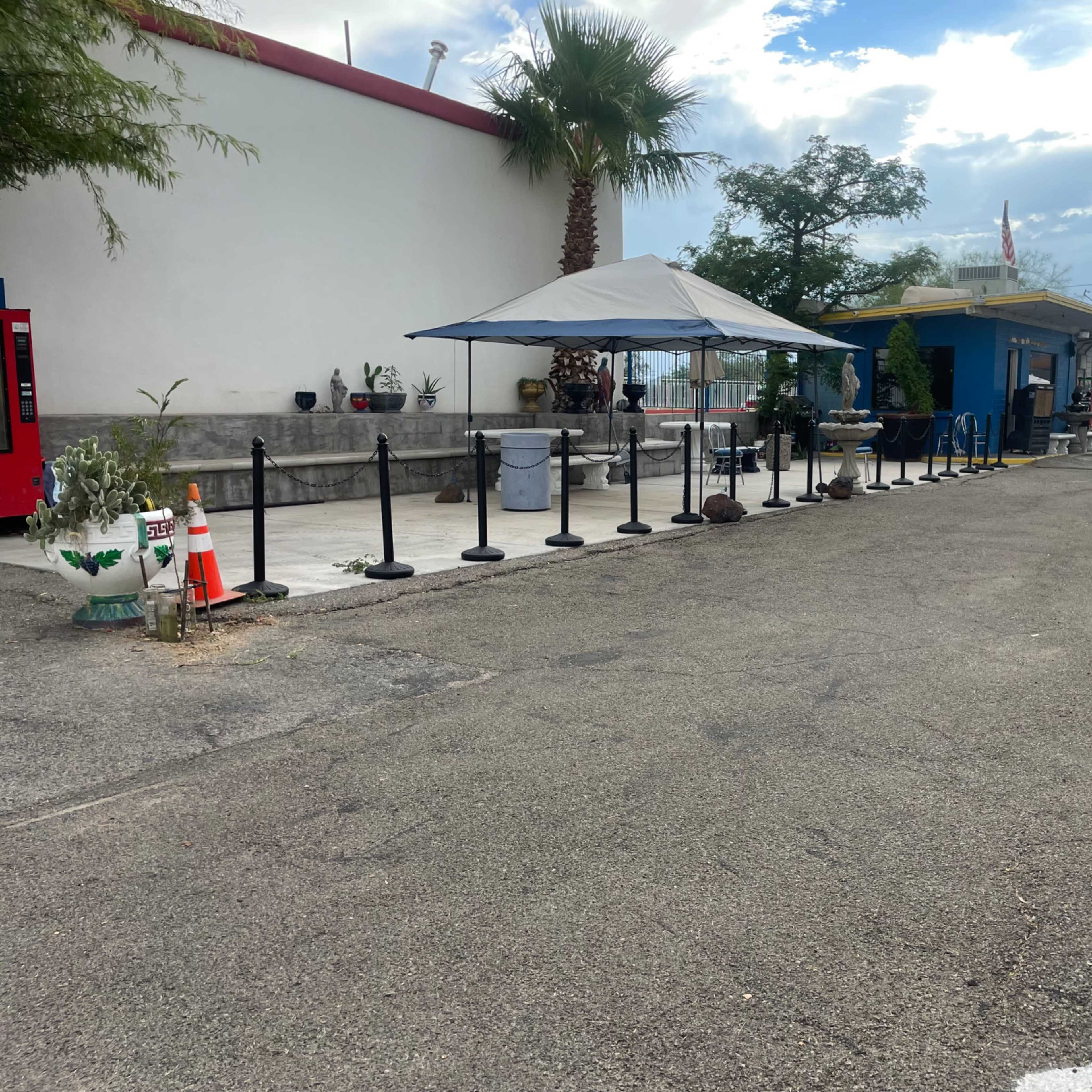 A paved parking area features a blue building with an awning, surrounded by various potted plants and a line of black stanchions.