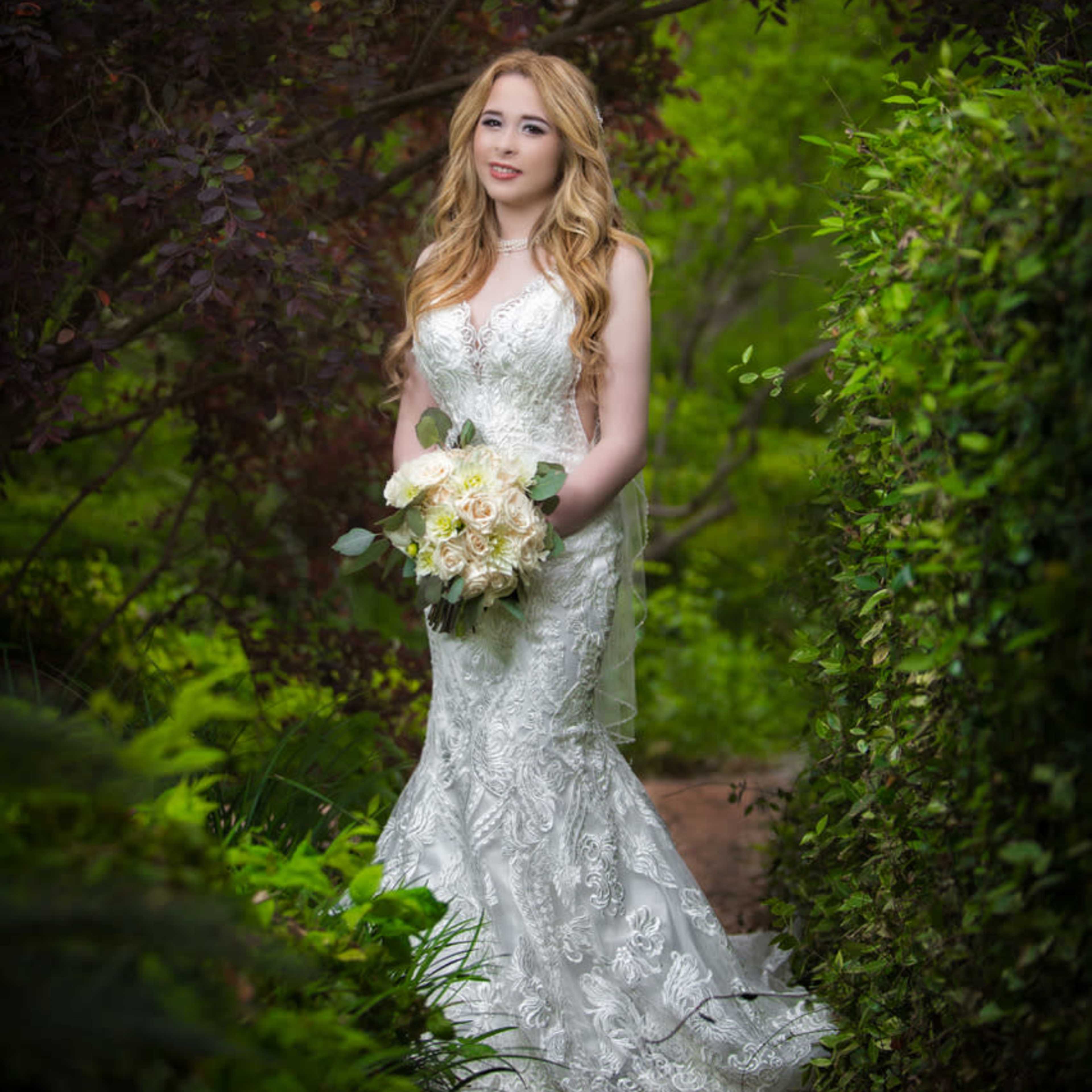 A woman in a white wedding dress stands gracefully in a lush green garden holding a bouquet of flowers.
