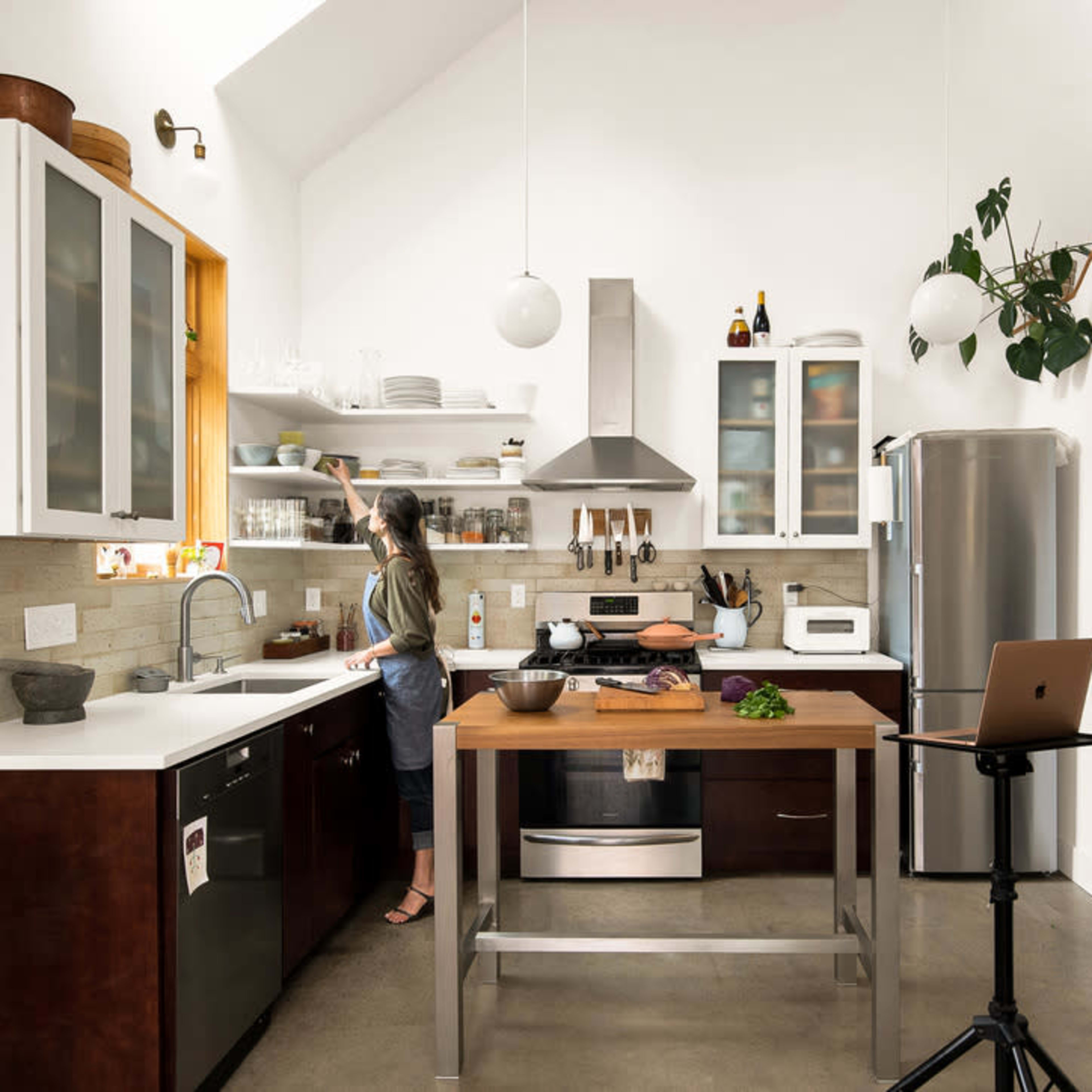 A woman stands at a modern kitchen counter, preparing food in a well-organized, brightly lit space featuring stainless steel appliances and open shelving.
