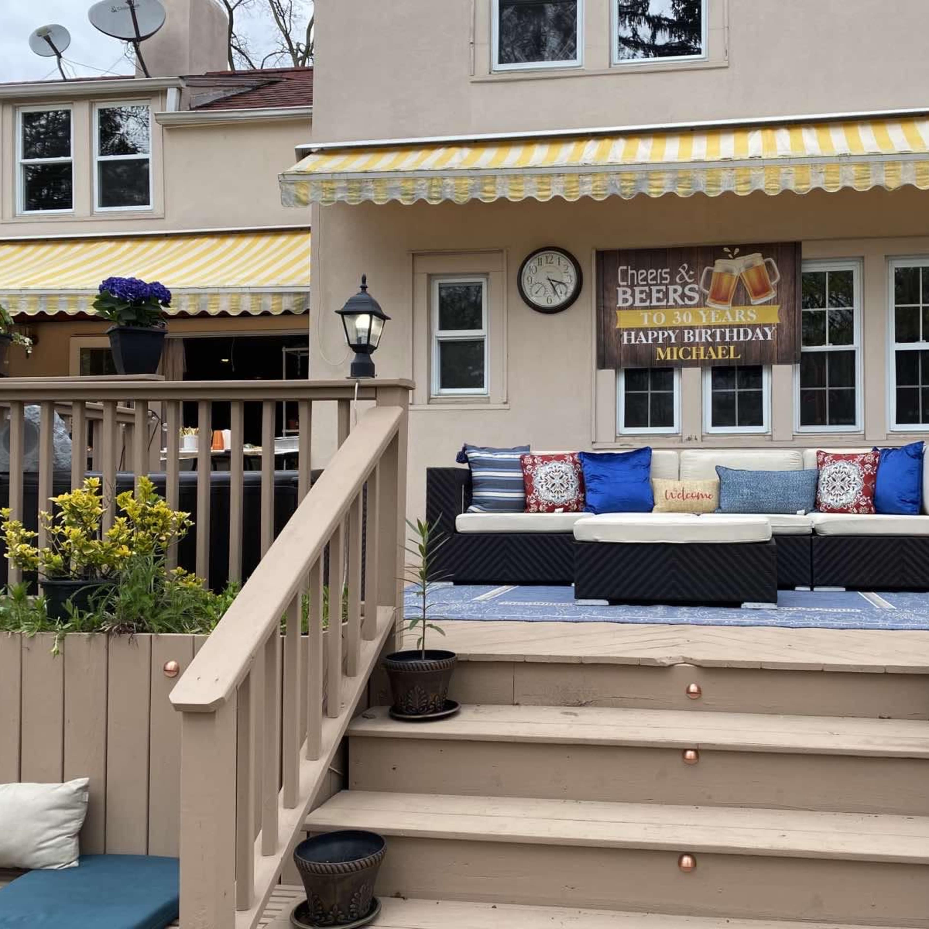 A birthday banner hangs above a patio area with a seating arrangement, flanked by flower pots and a clock on the wall of a house.