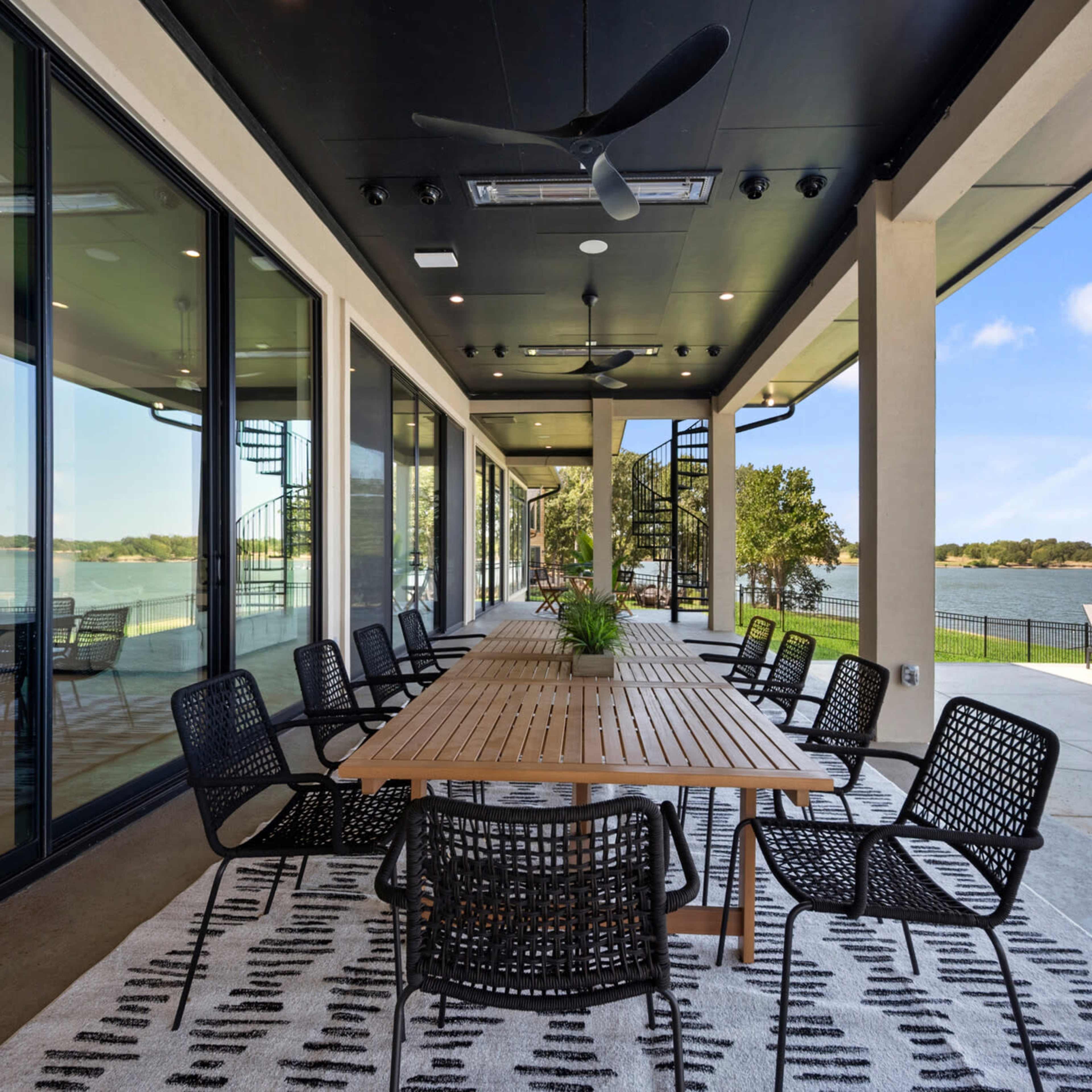 A spacious outdoor patio features a long wooden dining table surrounded by black chairs, overlooking a lake.