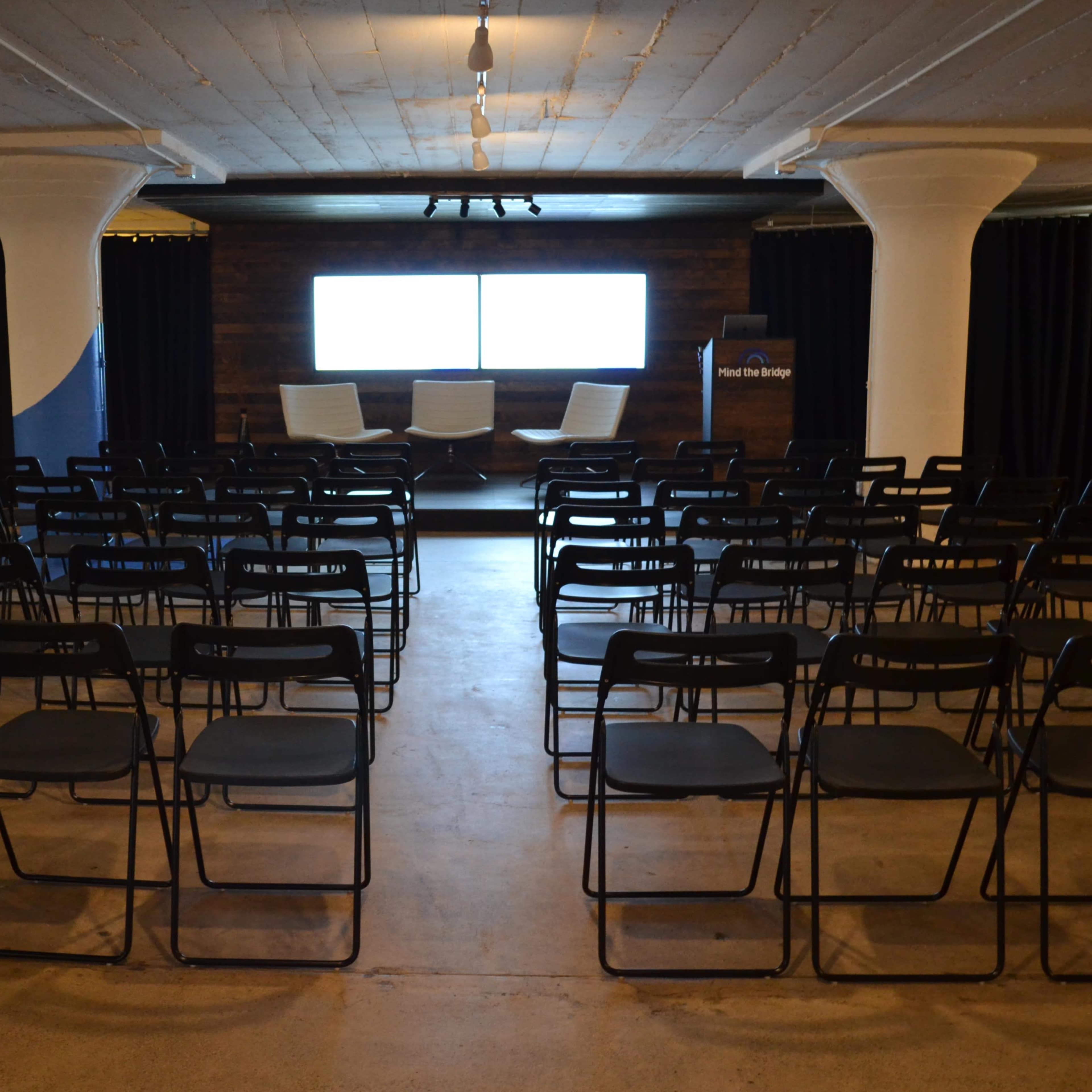 A conference room arranged with rows of black chairs facing a stage with three white chairs and a large screen.